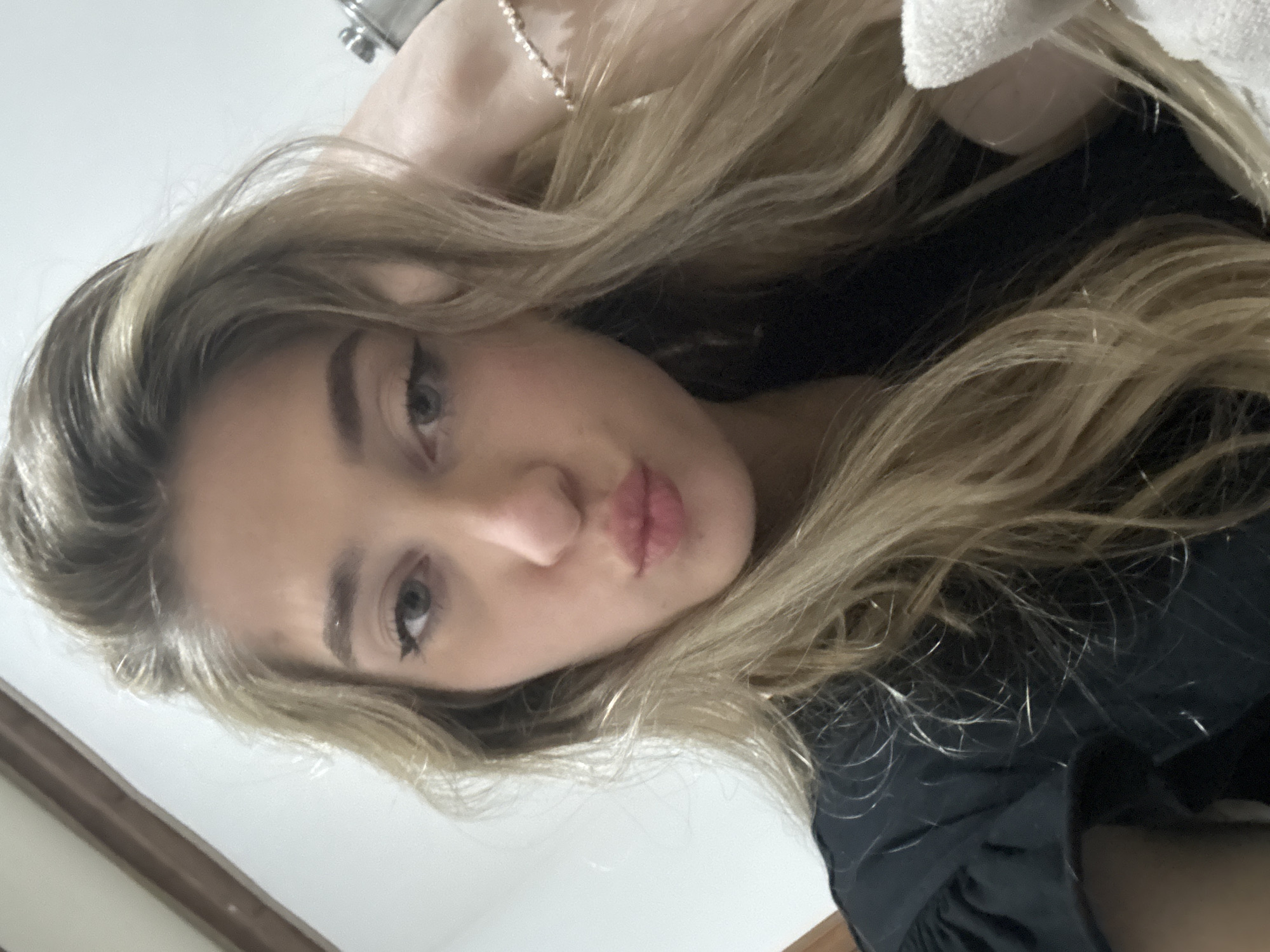 A young woman with blonde hair, wearing a black top, poses for a selfie. She has light eyes and her lips are puckered. Her hair is long and wavy, partially covering her face. The background is blurry and indeterminate, suggesting an indoor location. The city is Unknown, Unknown.