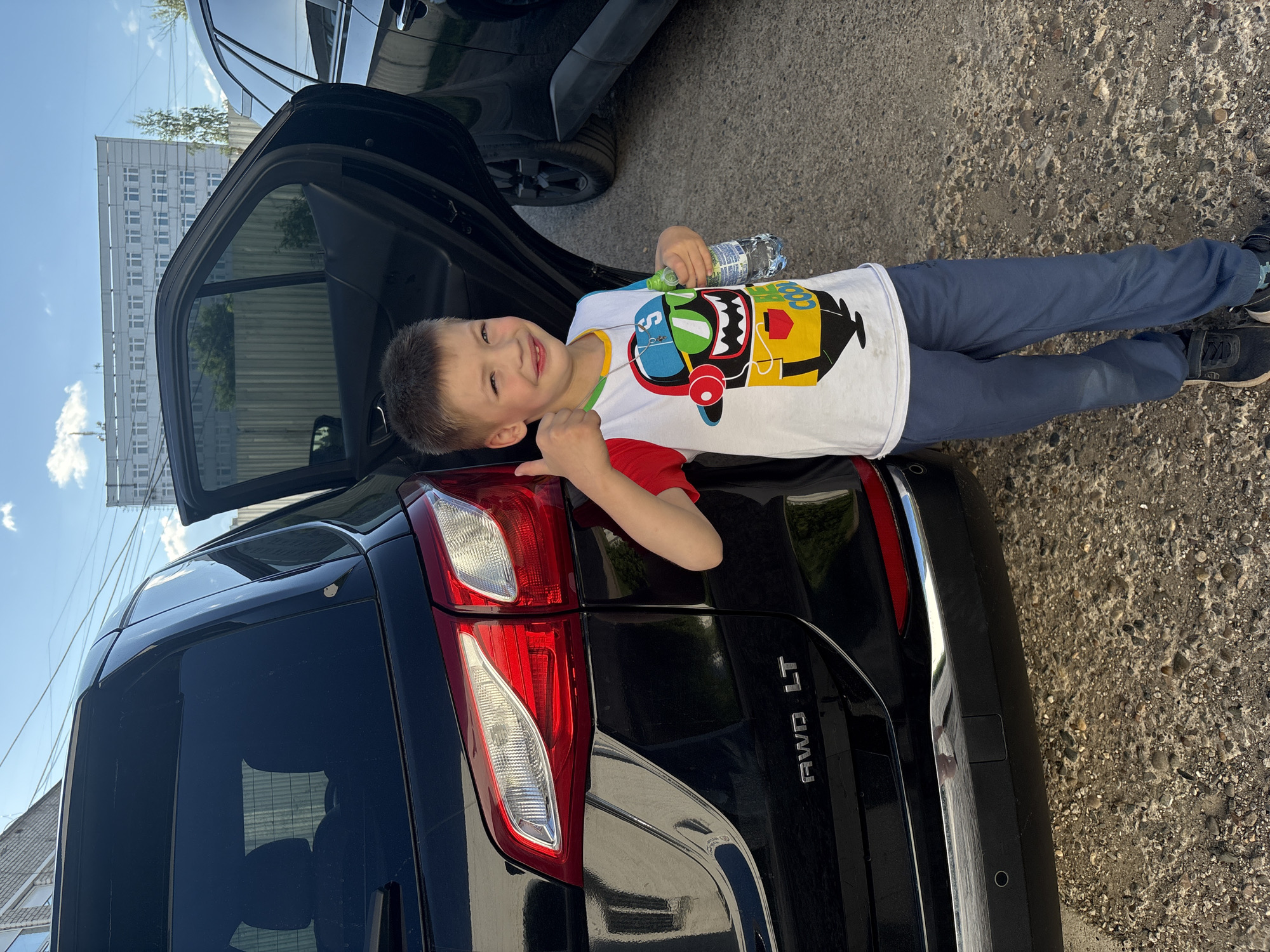 A young Caucasian boy, approximately six to eight years old, stands with his left side towards a black SUV. He is wearing a white t-shirt with a colorful graphic of a cartoon character, blue pants, and dark sneakers. The boy is smiling and giving a thumbs-up gesture with his left hand. He is holding a clear plastic bottle of water in his right hand. The SUV has its rear passenger door open. In the background, a wooden fence and a multi-story building with many windows are visible, suggesting an urban environment. The ground is covered with gravel and asphalt. The overall scene appears to be outdoors during the daytime, with a partly cloudy sky. This scene is located in the city of Chaikovsky, Russia.