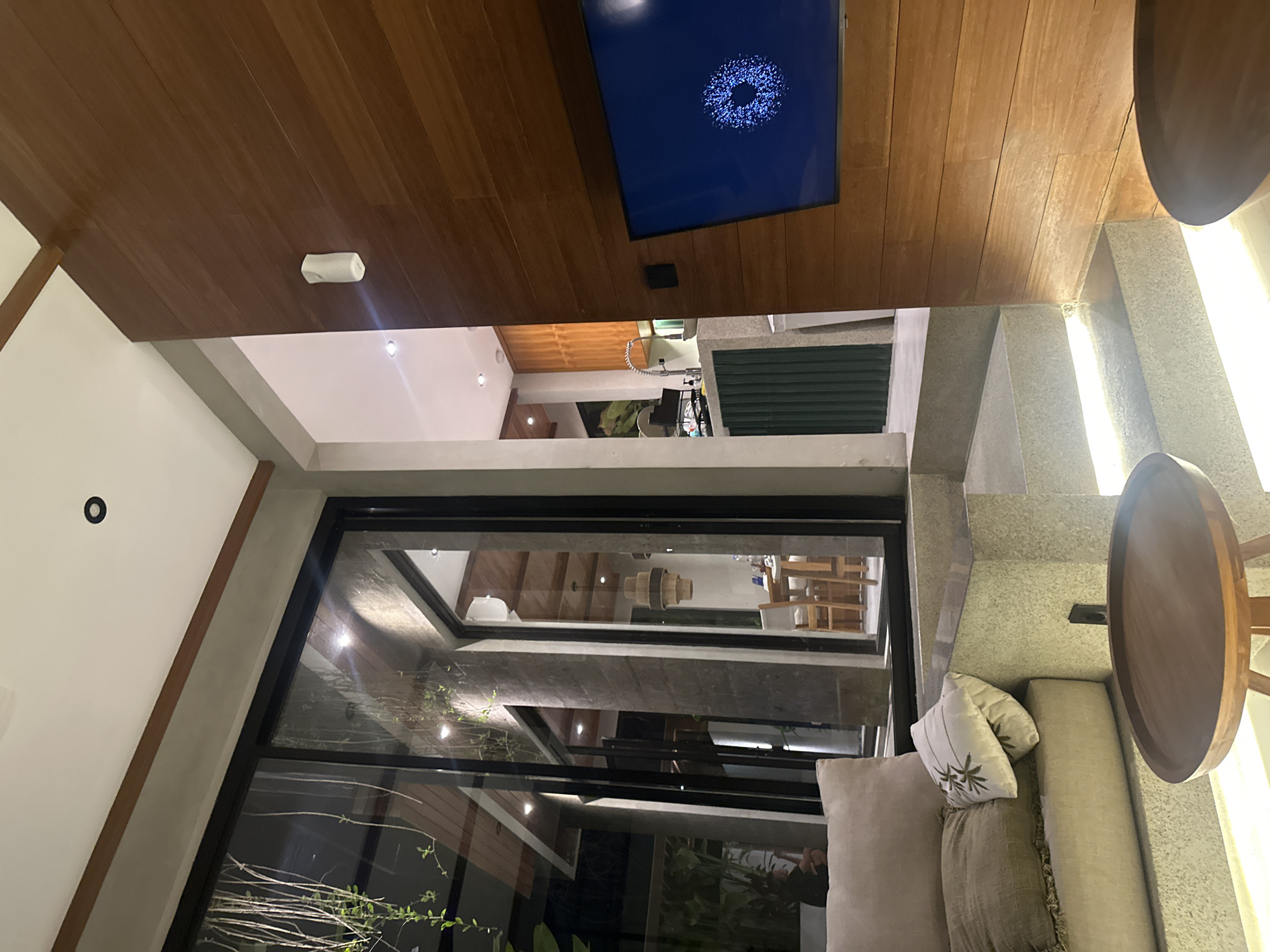 The image shows an interior space in Tumbak Bayuh, Indonesia. A large window offers a view of an outdoor area with plants. Inside, there's a seating area with a light-colored sofa and decorative pillows. A round wooden table stands nearby. The ceiling is constructed of wood planks, and a television screen is mounted on it.