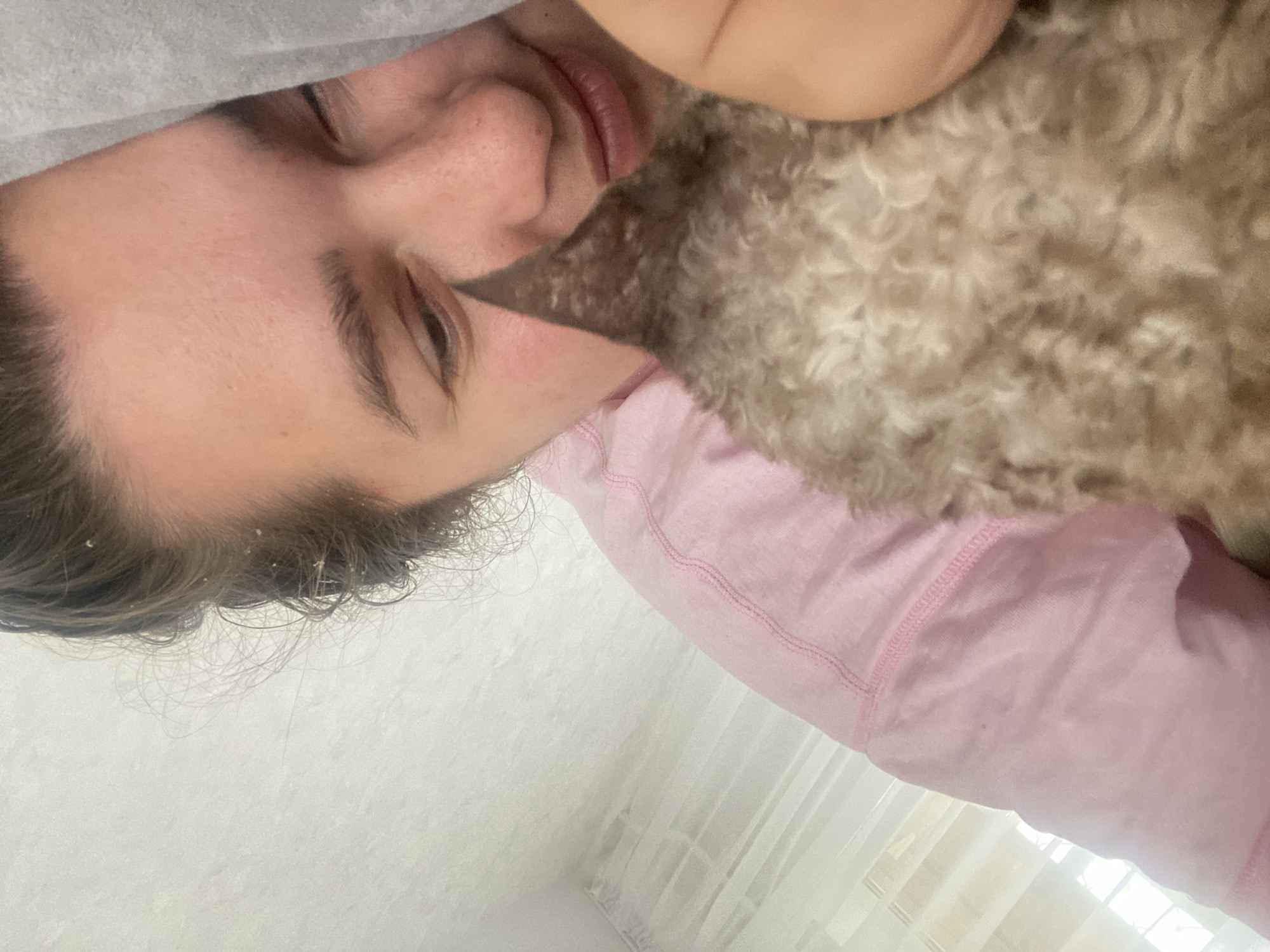An individual is lying in bed, partially obscured by a fluffy, light brown dog that is resting its head against the individual's face. The individual is wearing a pink long-sleeved shirt. The background includes white curtains and a white wall. The image appears to be a close-up selfie taken indoors, possibly in a bedroom. There are no indications of specific city locations.