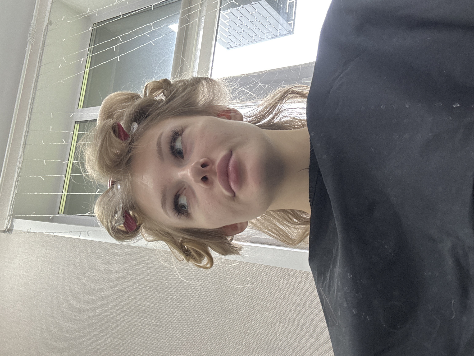 The image shows a young woman with blonde hair in rollers, looking upwards and to the right. She is wearing a black cape. The background includes a textured wall and a window with string lights. The scene appears to be an indoor setting, possibly a hair salon. The location is not specified within the image.