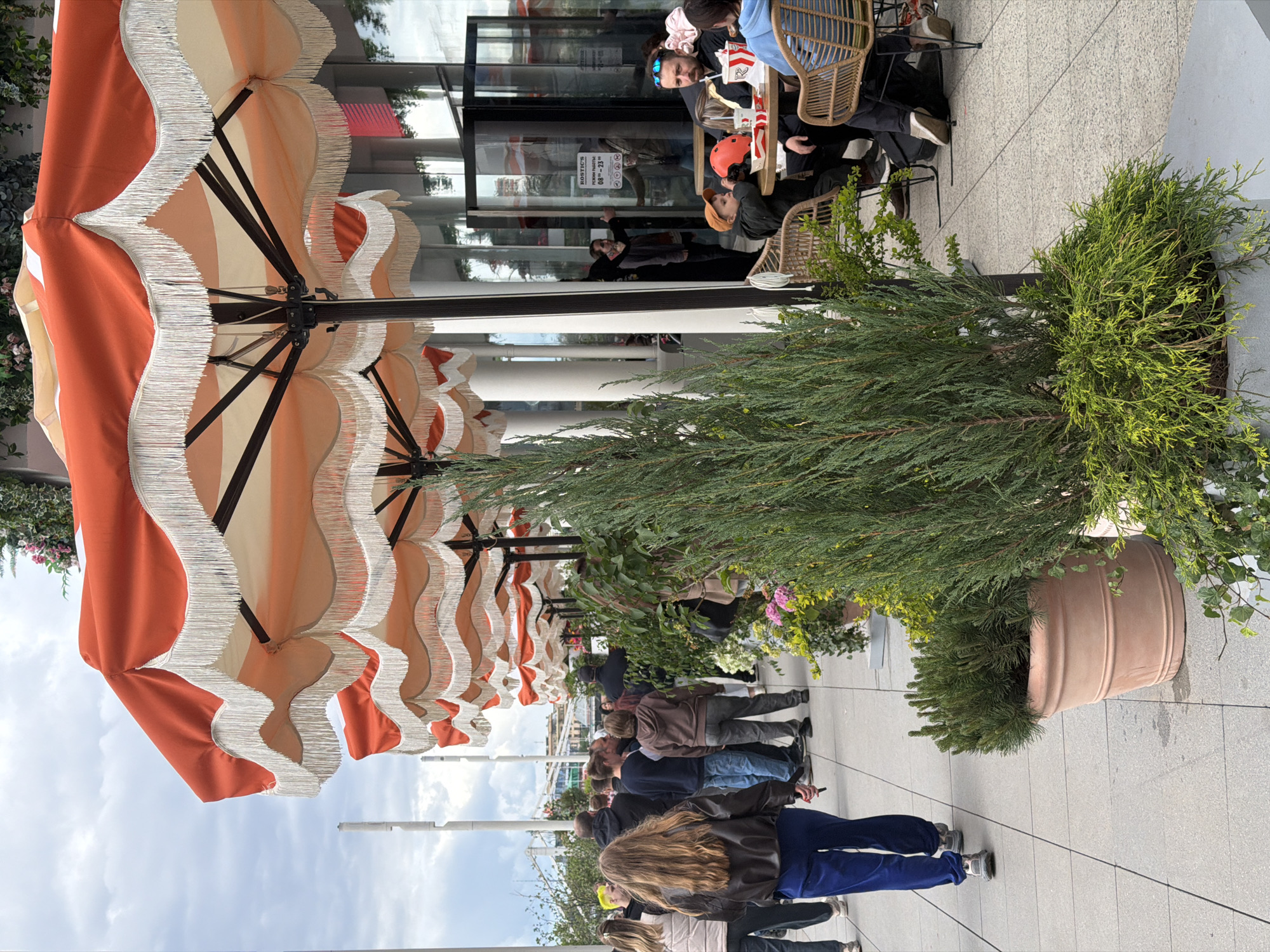 The image shows an outdoor scene likely in Moscow, Russia, based on the Cyrillic text visible on a storefront. A row of orange and white striped patio umbrellas with white fringe provides shade for an outdoor dining area. Patrons are seated at wicker chairs around tables outside a building with glass doors and windows, with people visible inside. A large potted evergreen plant sits beside the tables and umbrellas. A line of people is partially visible further down the sidewalk. The sky is overcast. The scene suggests a restaurant or cafe setting.
