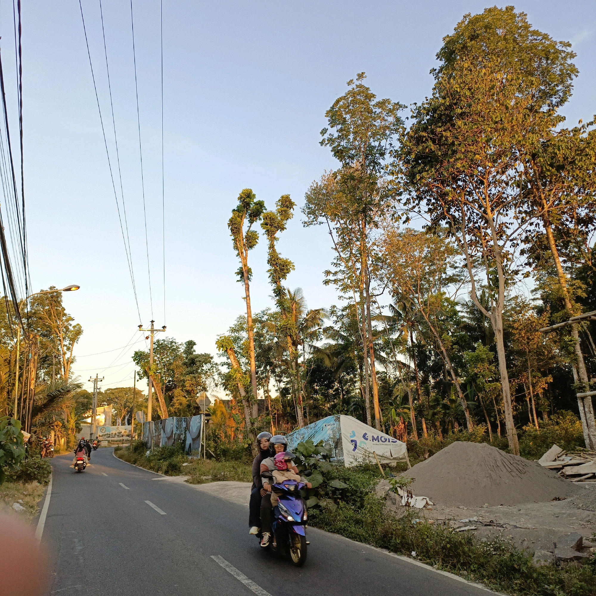 This is a photograph taken at sunset on a paved road in Salatiga, Indonesia, likely in a semi-rural or suburban area given the presence of tall trees and a pile of construction sand. The scene depicts a street with several motorcycles. In the foreground, a motorcycle is carrying three people: a driver wearing a helmet, a passenger behind the driver also wearing a helmet, and a child sitting between them, also with a pink helmet. The motorcycle is dark blue and appears to be in motion. Further down the road, several other motorcycles with riders are visible, also moving away from the camera. The road has dashed white lines marking the lanes. On the right side of the road, there is a large pile of sand and some construction materials, including wooden planks. A large white tarp with text, possibly a banner or advertisement, is propped up behind the sand pile. The left side of the road features a dense line of tall trees and vegetation, with some utility poles and overhead power lines. The sky is clear and a pale blue, illuminated by the warm light of the setting sun, which casts long shadows and highlights the tops of the trees with golden hues.