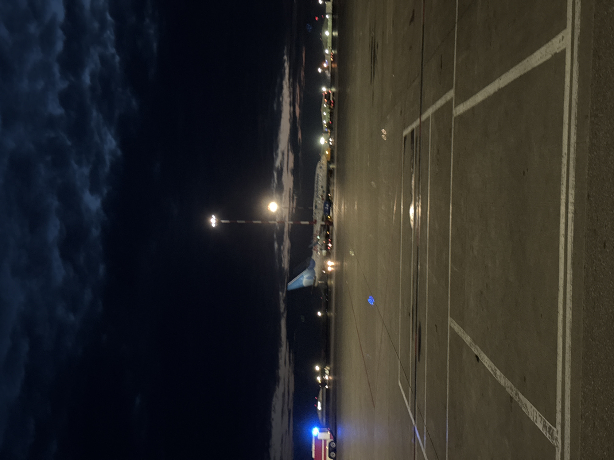 At night on an airport tarmac in Khimki, Russia, a fire truck with flashing blue lights is positioned to the left of a passenger airplane. The airplane, bearing the airline's livery, is stationary on the runway, illuminated by various lights. Other airport vehicles and lighting structures are visible in the background. The sky above is dark and cloudy. The foreground shows the markings on the tarmac, indicating parking or taxiing areas.