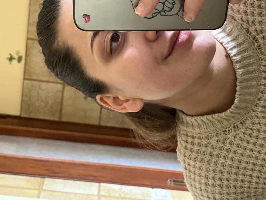 The image depicts a partial reflection of a female individual taking a mirror selfie in Ancenis, France. The subject's face is visible from the forehead to the chin, featuring light brown eyes and a subtle smile. Her dark hair is pulled back. She wears a light-colored, textured knit sweater. Her right ear is visible. She holds a gray smartphone vertically, obscuring the right portion of her face. The smartphone screen is cracked, displaying a dark pink, possibly heart-shaped, sticker in the upper right corner and a reflected outline resembling a skeletal hand. The background, visible in the mirror, consists of light beige rectangular wall tiles arranged in a staggered pattern. To the left, a brown wooden vertical frame, consistent with a window or door, is present. A faint dark green, indistinct shape is reflected on the tiled wall above the subject's head. The scene is illuminated by ambient light.