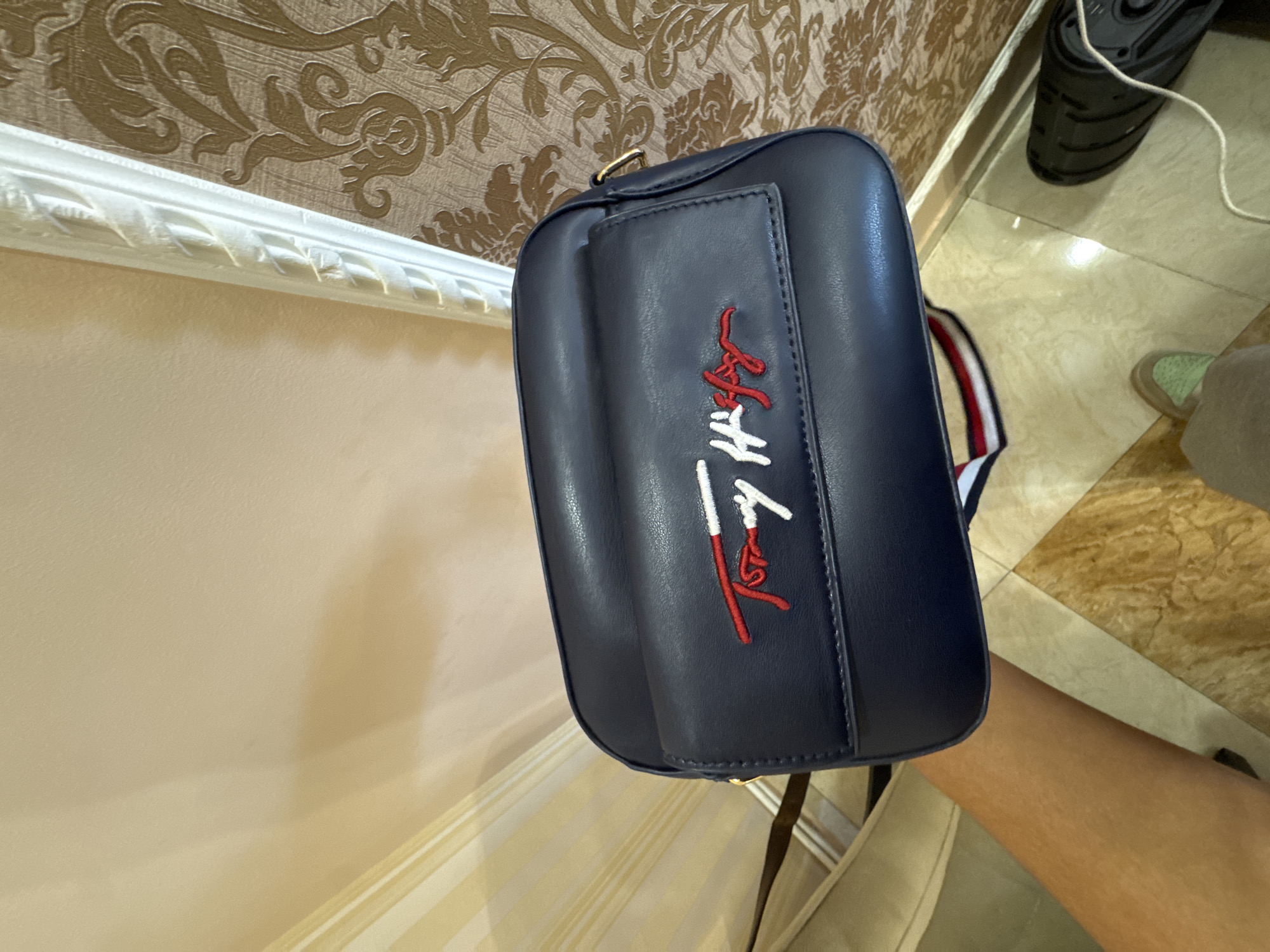 A close-up shows a blue "Tommy Hilfiger" brand crossbody bag being held up against a wall with decorative trim. The bag features the brand's logo embroidered in red and white. Part of a person's arm is visible, as well as a bit of flooring. The background includes a wall with ornamental wallpaper and a black shoe and floor. The location is Yerevan, Armenia.