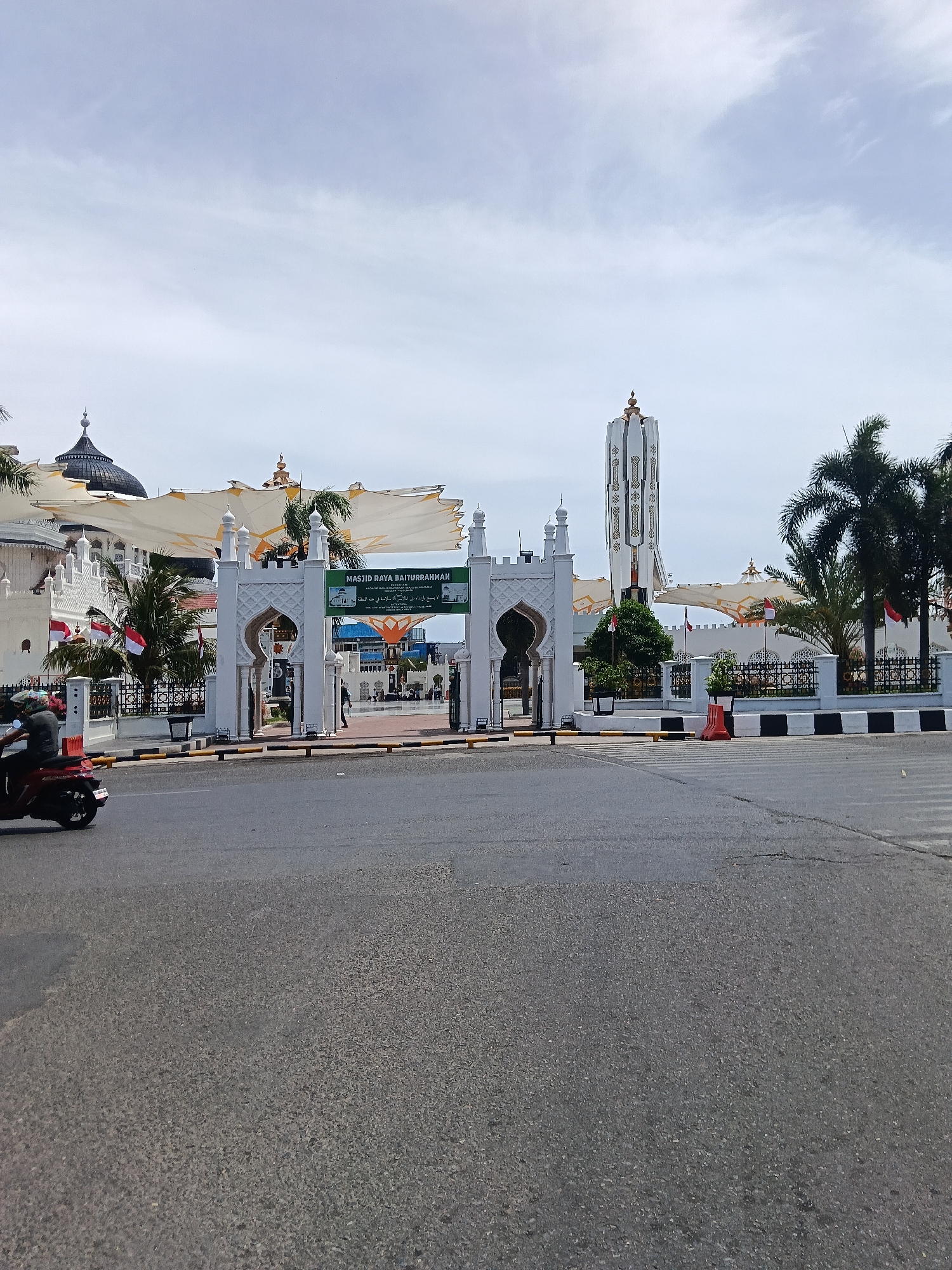 The image shows the entrance to the Baiturrahman Grand Mosque in Banda Aceh, Indonesia. A white, ornate gate with Islamic architectural elements stands at the entrance, with a banner displaying "MASJID RAYA BAITURRAHMAN" in green and white. Beyond the gate, the mosque's white walls, traditional roof structures covered by large white awnings, and a tall, slender minaret are visible. Indonesian flags are displayed in front of the mosque. The scene is set on a street with an asphalt road in the foreground, where a person on a red scooter is riding towards the left. The sky is cloudy and bright, indicating daylight. The overall environment suggests a public space in a city.