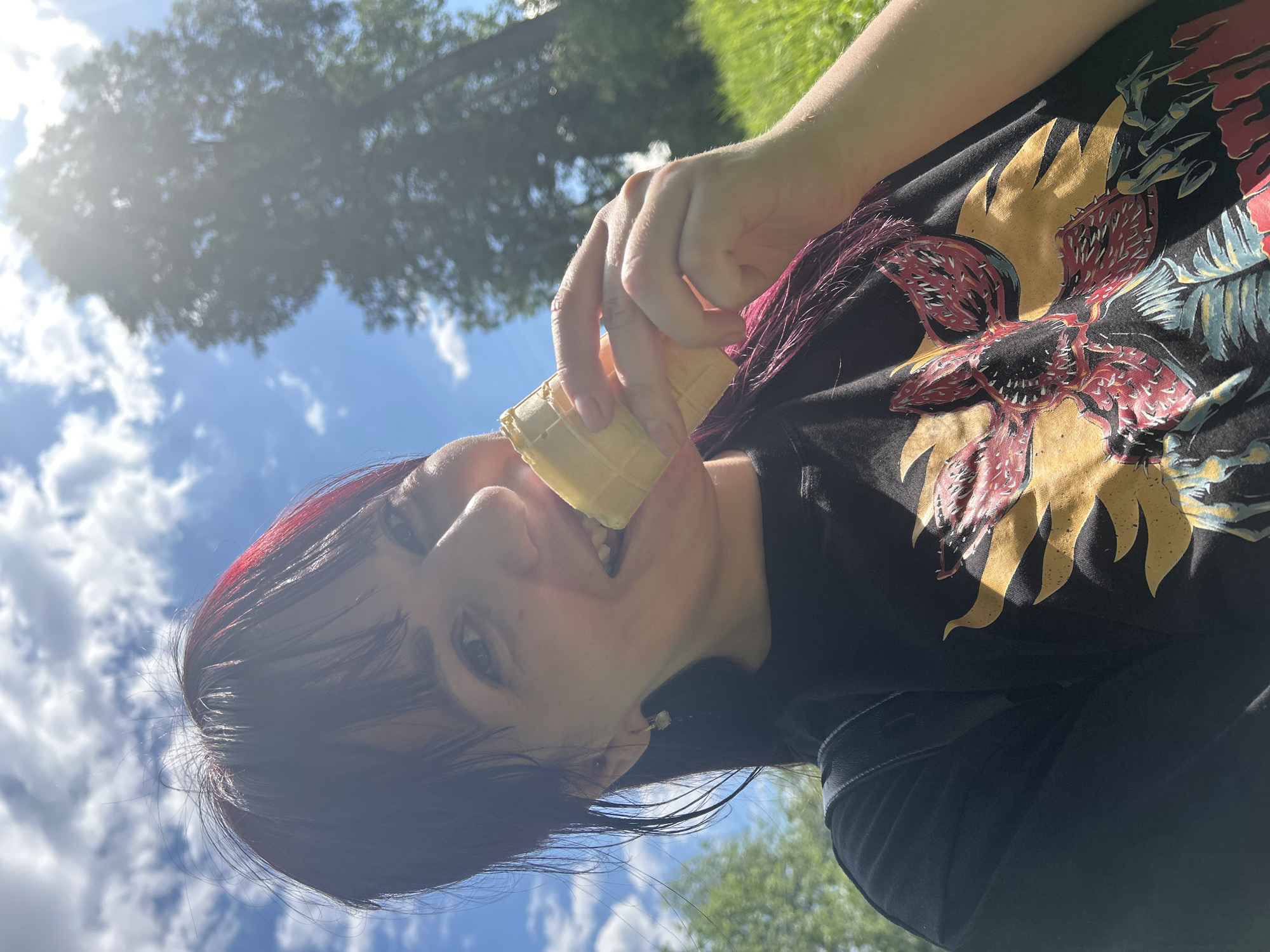 A young woman with dark hair and red streaks is captured in a close-up selfie, holding a wafer cone to her mouth. She is outdoors, with a bright blue sky and scattered clouds in the background. Sunlight filters through the leaves of a large tree on the right side of the frame, casting dappled shadows across the scene. The woman is wearing a black t-shirt with a graphic print of a creature with red and yellow elements, possibly from a TV show or movie. She has a smile on her face, and her eyes are looking towards the camera. The grass is green and lush, suggesting a park or natural setting. The image has a casual, summery feel. The location is identified as Foky, Russia.