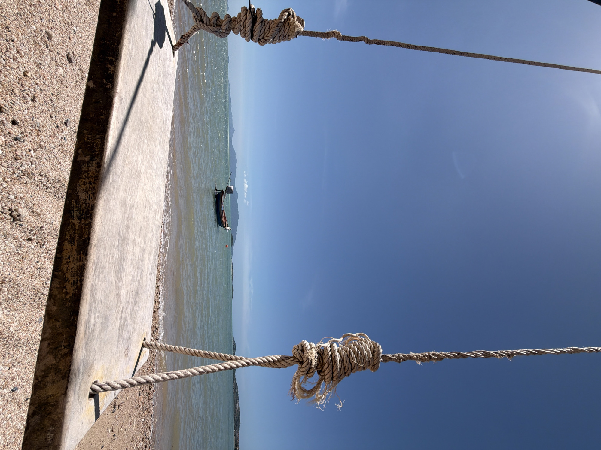 A low-angle, eye-level shot captures a swing at the beach in Ko Samui District, Thailand. The swing's wooden seat, weathered and worn, rests on a sandy beach composed of small pebbles. Two thick, braided ropes, each knotted securely to the seat, ascend into a clear, bright blue sky. The ropes are made of natural fibers and appear aged. In the middle distance, the turquoise ocean meets the horizon, with gentle waves lapping at the shore. A small, dark boat with an outboard motor is anchored in the water, and a few distant islands are visible as faint silhouettes against the sky. The sun casts a long shadow of the swing's support across the seat and onto the sand. The scene is devoid of people.