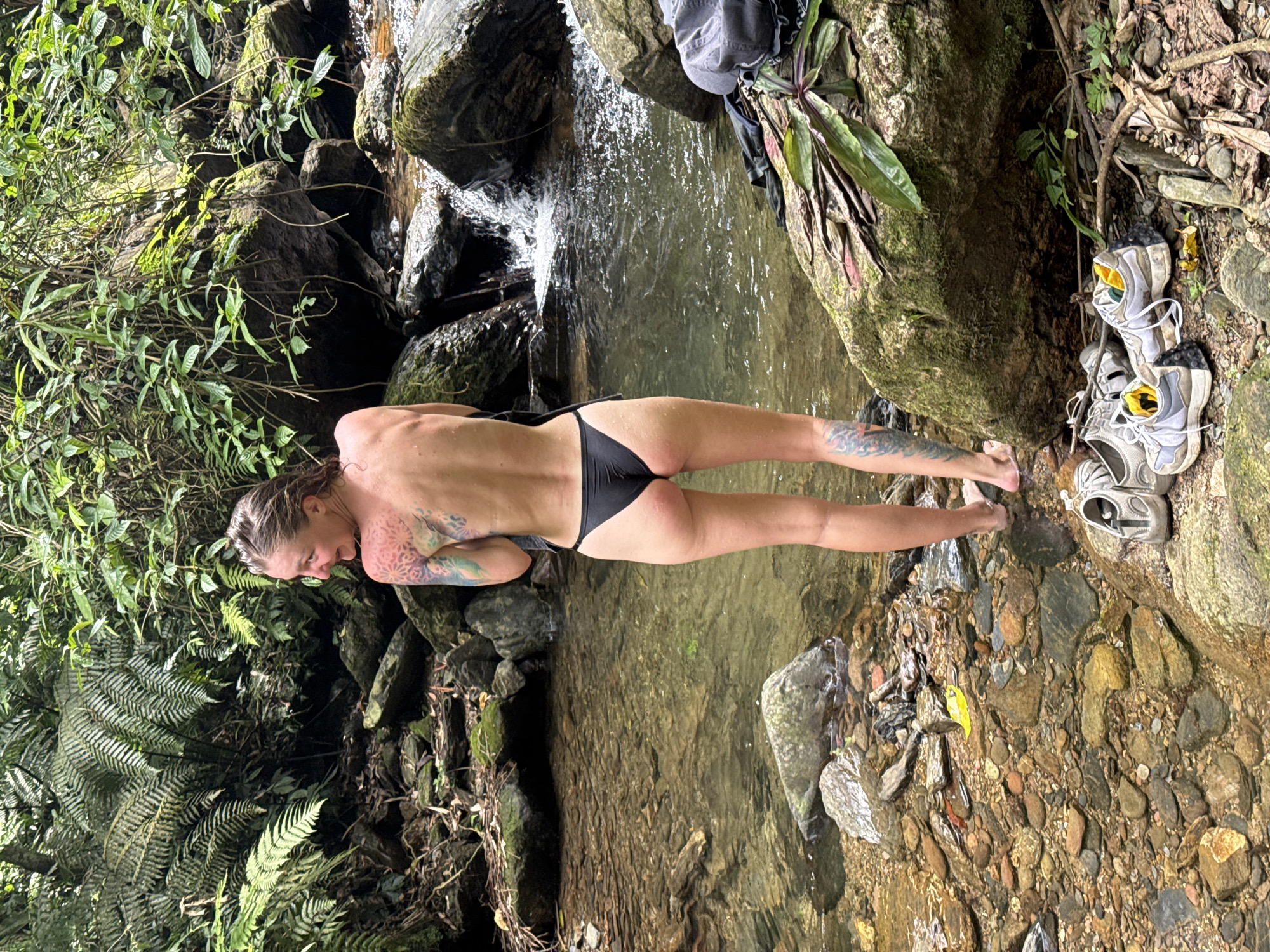 A nude woman in a black bikini bottom stands in a stream with her back to the viewer. She has tattoos on her arms and back. The stream appears to be at the bottom of a waterfall. Clothing and shoes are piled on a rock to the right of the woman. Ferns and other foliage are visible along the sides of the stream. The setting is reminiscent of a natural environment, potentially near Suối Giàng, Vietnam.