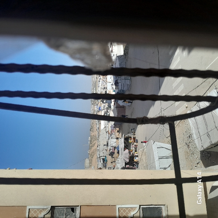 A view from a barred window reveals a landscape with a clear blue sky. Below, a sprawling makeshift settlement stretches across the arid terrain. Tents, dwellings, and market stalls are densely packed, indicating a crowded environment. A road with crosswalk markings cuts through the scene. The window frame is visible in the foreground, casting shadows on a nearby building.