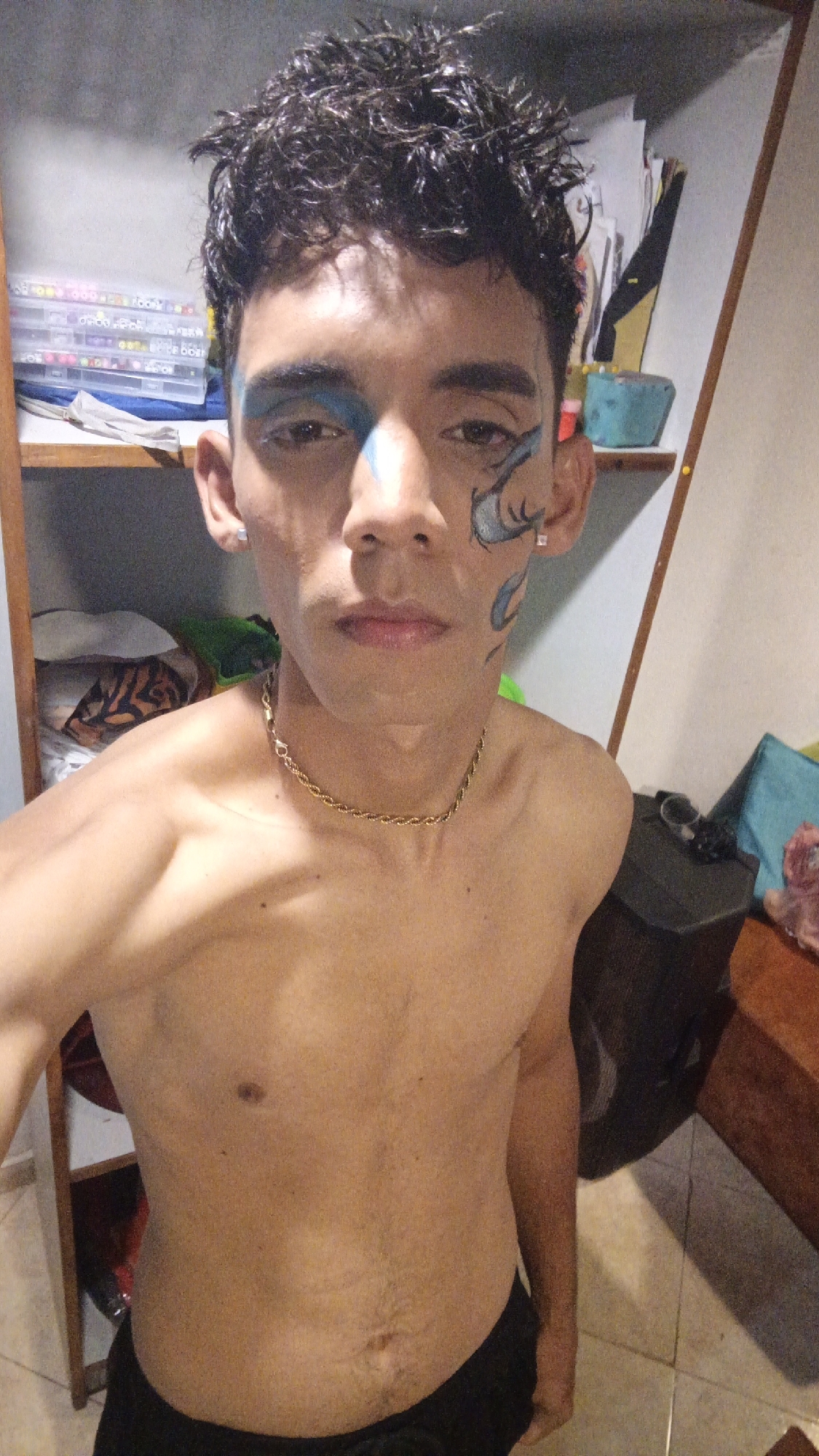 A shirtless young man with dark curly hair and face paint stands indoors, possibly in Playa El Agua, Venezuela. He is wearing a gold chain necklace and black shorts. His face paint consists of a blue stripe down the center of his forehead and a blue and white design on his left cheek. A shelving unit is visible behind him to the left, containing various items, including containers of beads. To his right is a dark speaker and a wooden table. The floor is tiled.