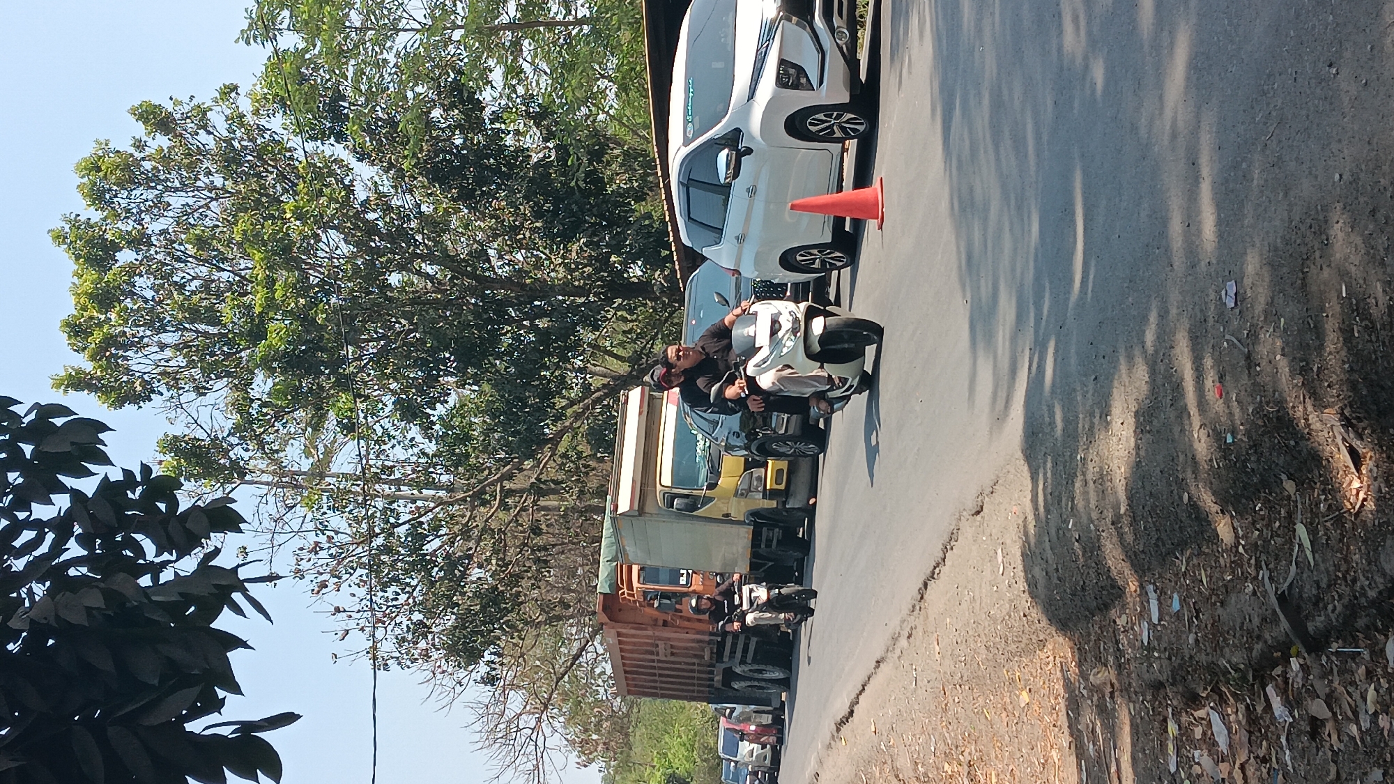 In Jonggol, Indonesia, on a sunny day with clear blue skies, a traffic jam is depicted on a paved road. The road is lined with trees on both sides and shows signs of wear and tear, with debris scattered along the shoulder. In the foreground, a white scooter carries two individuals; the driver wears a black shirt and sunglasses, while the passenger is dressed in a black polo shirt. To the left, another scooter, driven by a person in a black shirt, is also in motion. Behind these scooters, a yellow truck with a wooden cargo bed is stopped, followed by a black car. Further down the road, an orange truck is visible, and a white MPV is parked on the right side, marked by an orange traffic cone. The scene suggests a typical day with moderate traffic flow in a semi-urban or rural Indonesian setting.
