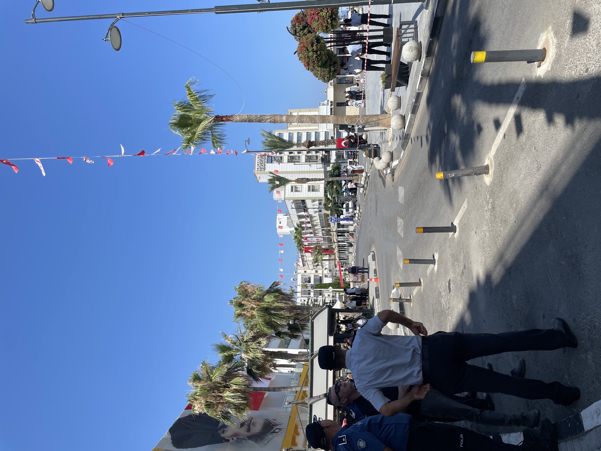 The image depicts a street scene in Kyrenia, Northern Cyprus. In the foreground, three men in police uniforms are standing and conversing. One man on the left wears a dark blue uniform with a badge and a cap, while the man in the center wears a light blue shirt and dark trousers, also with a cap. The third man on the right wears a similar dark blue uniform and cap. Behind them, other individuals are visible, some appearing to be civilians.
The street is paved, with bollards and a sidewalk present. Palm trees are scattered throughout the scene, and buildings line the street in the background. Flags, primarily red and white, are displayed on poles and strung across the street, suggesting a celebratory or national event. One large banner on a building features a portrait, possibly of a national figure. The sky is clear and blue, indicating a sunny day. The overall impression is a public space with a police presence, possibly during an organized event or gathering.