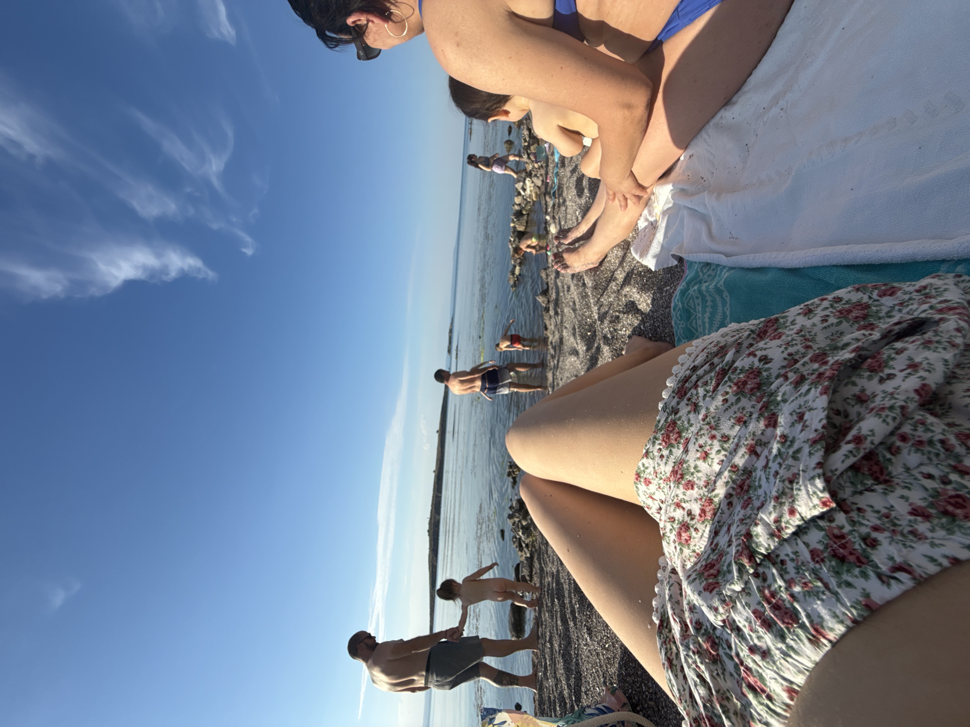 The image depicts a sunny day at a beach in Sozopol, Bulgaria. Several people are present, enjoying the water and the shore. In the foreground, a person wearing a floral skirt is partially visible. Further back, a man in swim trunks stands by the water, holding hands with a child who is nude. Another man in swim trunks is further out in the water. In the background, more people are scattered along the rocky shoreline. The sky is clear blue with some wispy clouds. The sun appears to be high in the sky, suggesting it is midday or early afternoon. The overall mood seems relaxed and summery. No visible text is present in the image.