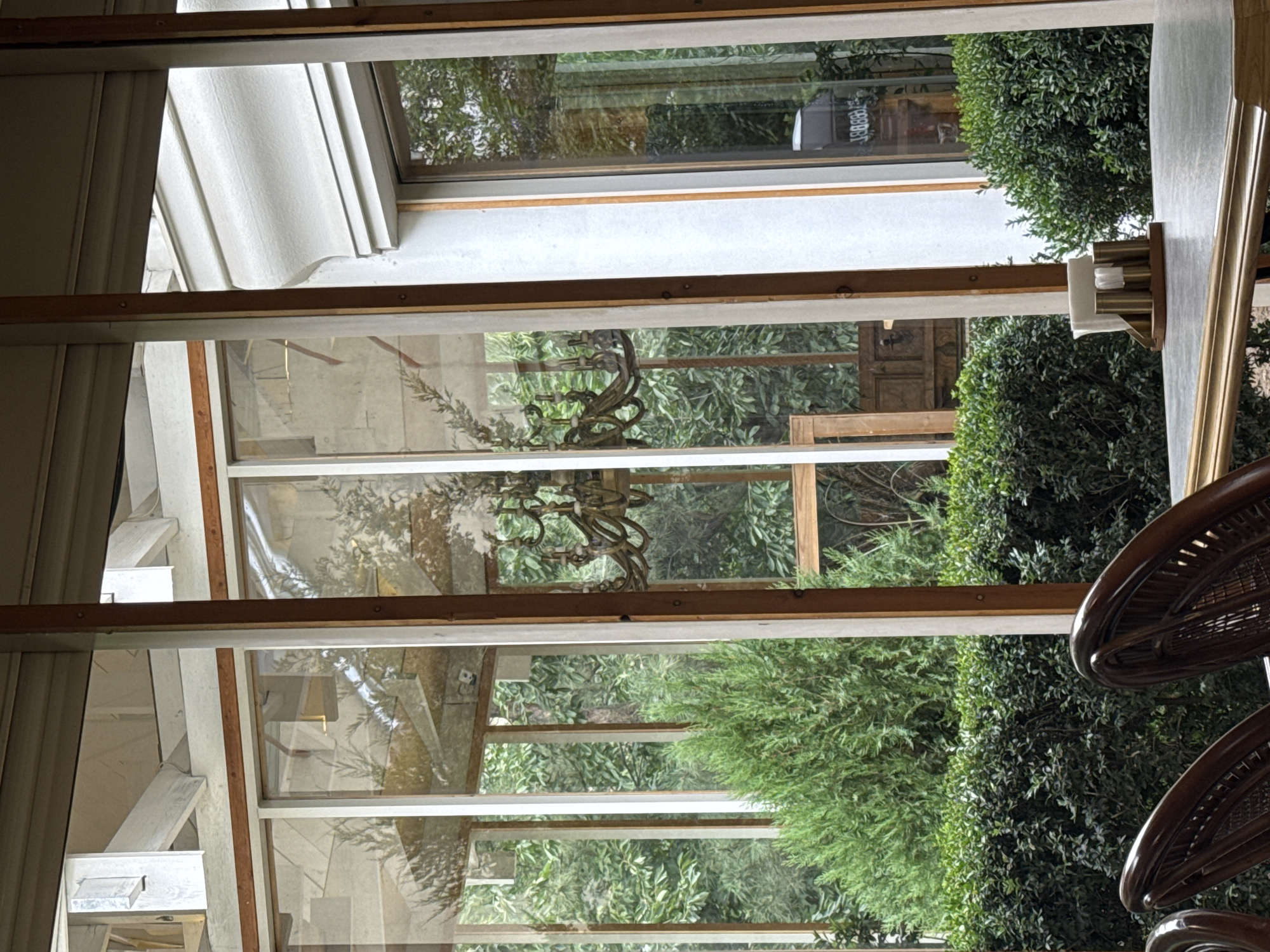 The image shows an interior view through a large glass-paned wall, possibly a sunroom or conservatory, looking out onto a lush green garden. In the foreground, parts of two dark wicker chairs are visible, along with a table with a light-colored, textured surface. On the table, a metallic condiment holder with napkins and shakers is positioned. Through the glass, a decorative brass chandelier with multiple arms hangs in the middle ground. The garden outside is dense with trees and shrubs, creating a natural backdrop. The architecture of the building is visible through the glass as well, with wooden beams and white framing. The overall scene suggests a tranquil setting, possibly a restaurant or a private residence, with a focus on natural light and greenery.