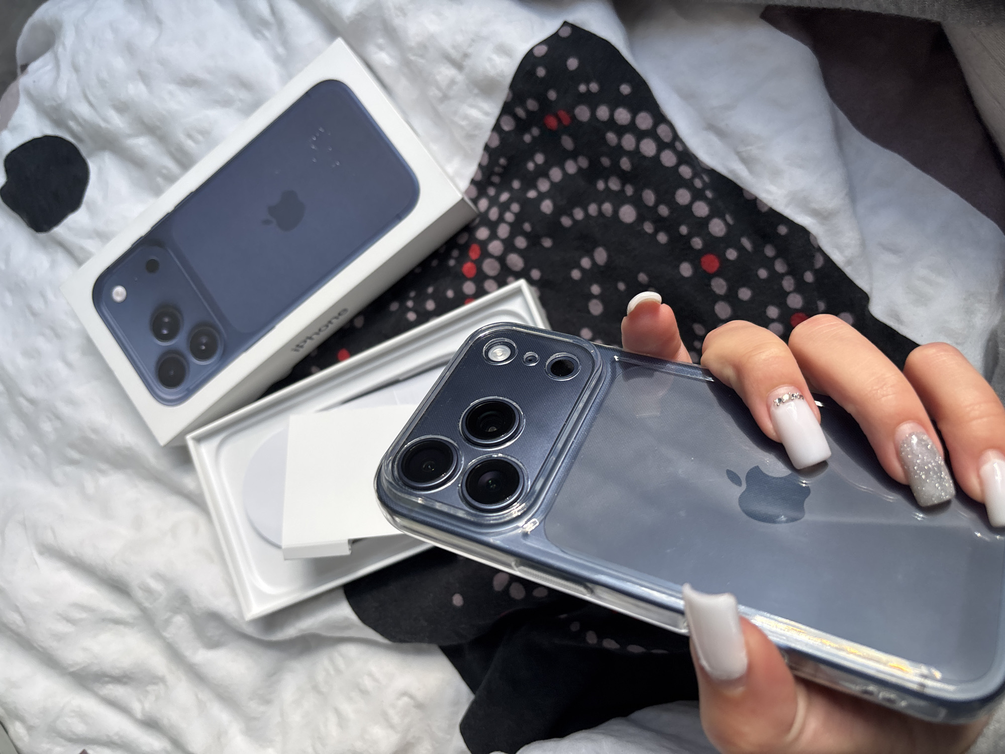 A hand is holding a blue iPhone in a clear case, with its original box placed on a patterned fabric. The environment is a bedroom. No evidence suggests that the image was taken in Gabrovo, Bulgaria.