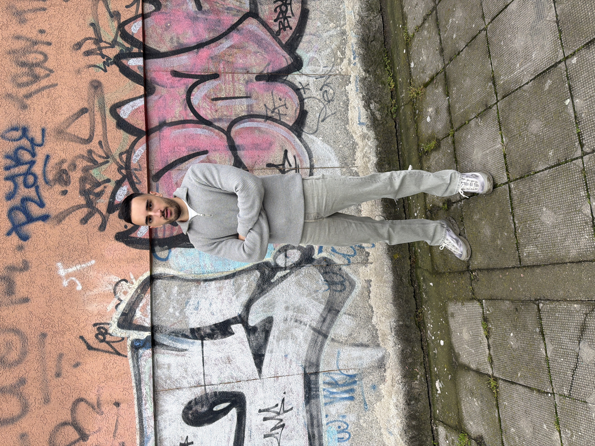 A man stands in front of a wall with graffiti. The wall is divided into two sections: the left side is orange with blue and black graffiti, while the right side is gray with pink and black graffiti. The man is wearing a gray ribbed sweater and matching pants. He has his arms crossed and is looking directly at the camera. The ground is paved with gray blocks with moss growing in the cracks. The location is possibly in Plovdiv, Bulgaria.