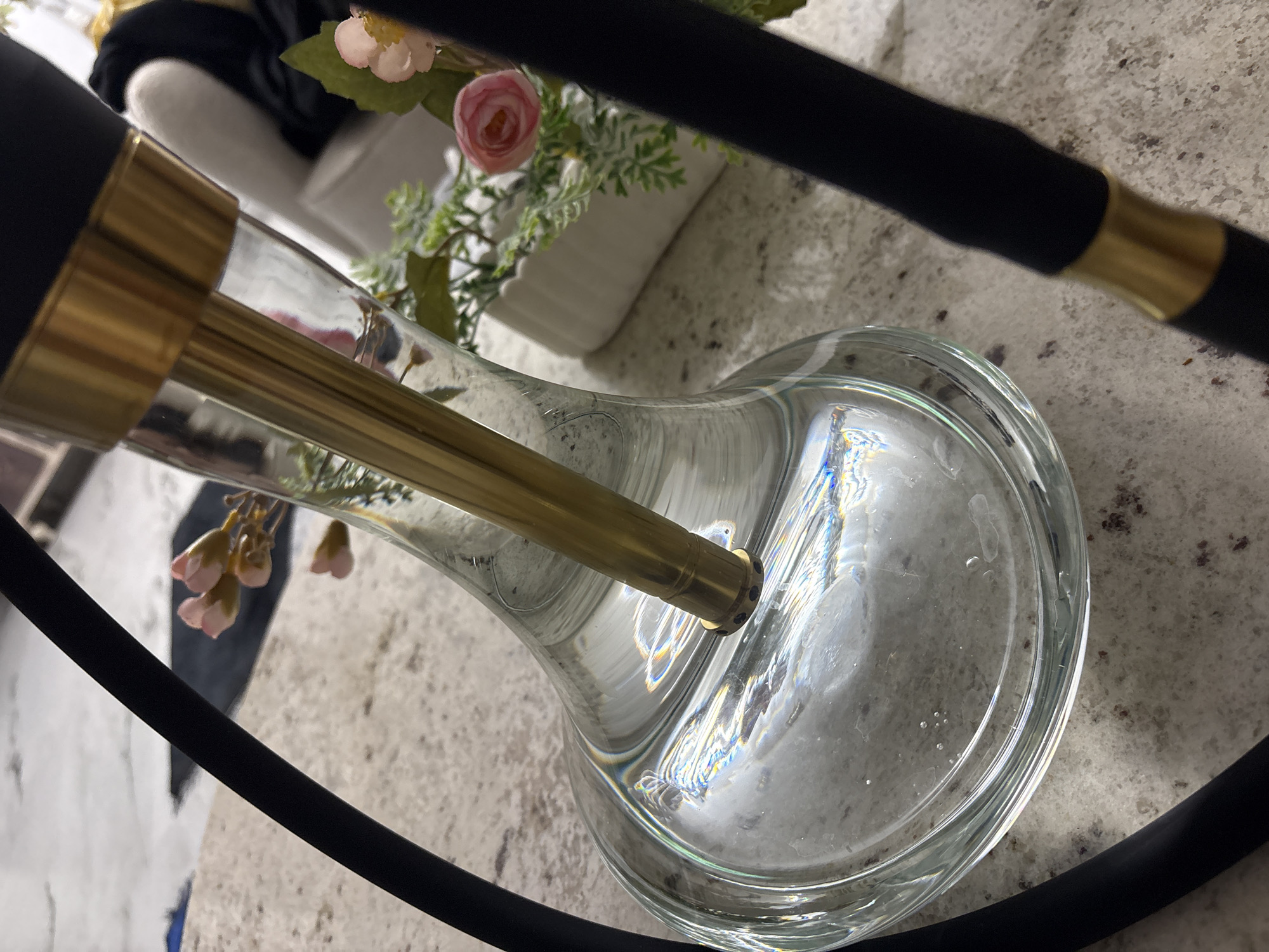 A close-up, eye-level shot captures a portion of a hookah with a clear glass base filled with water, a gold downstem, and a black hose. The water reflects light, creating a prismatic effect. In the background, a white vase with artificial pink flowers and greenery is visible on a speckled countertop. A light-colored upholstered chair and a dark fabric are also partially visible in the background, suggesting an indoor setting. The image is taken from a perspective looking down into the hookah base. There are no people or identifiable landmarks that would suggest a specific location within Kadewatta, Sri Lanka.