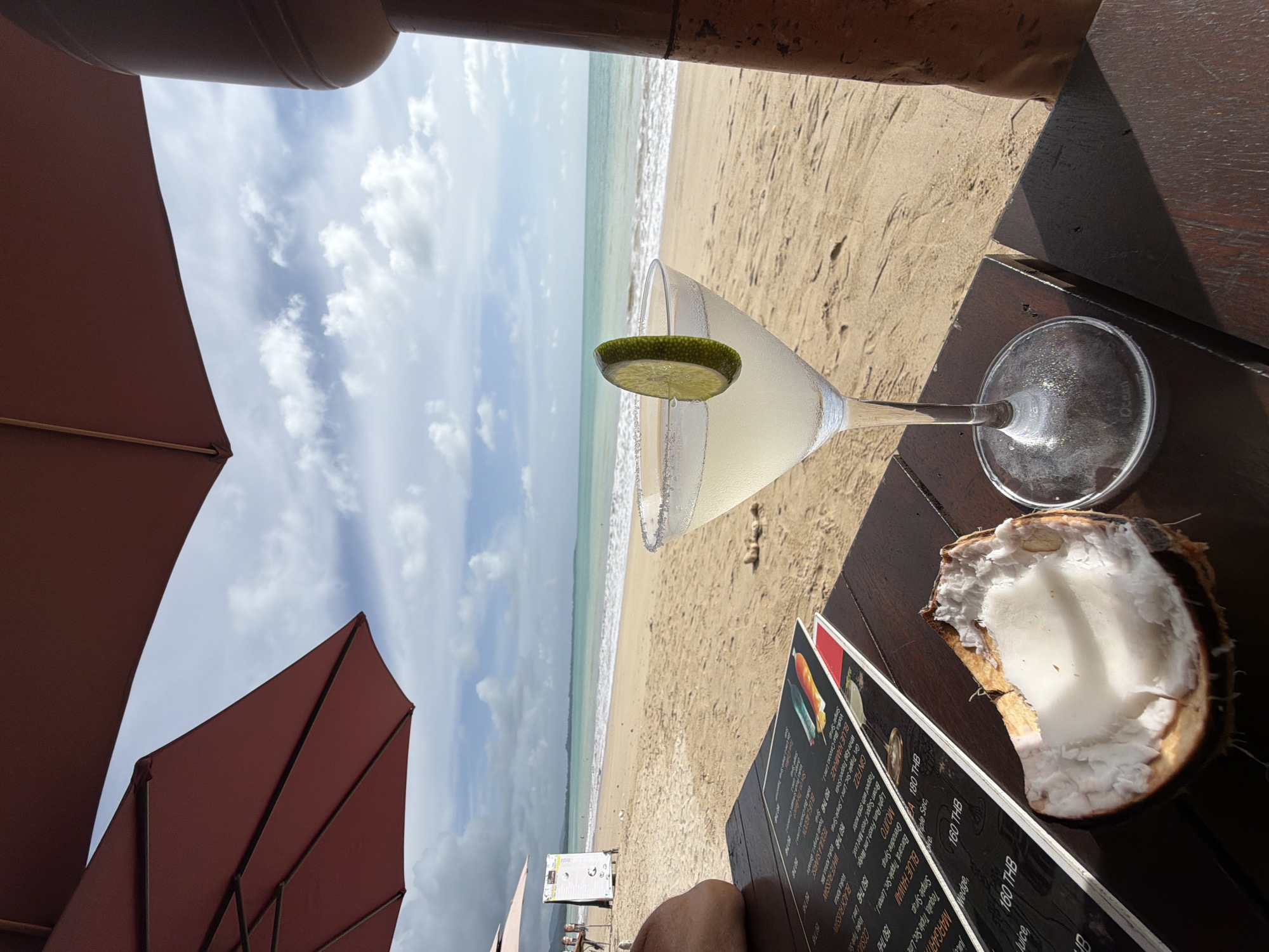 The image depicts a beachside dining scene in Bang Muang, Thailand, under overcast skies. In the foreground, a wooden table holds a martini glass filled with a pale yellow liquid, garnished with a lime slice and rimmed with salt. Adjacent to the glass is a cracked-open coconut with its white flesh exposed. A menu, with some pages appearing glossy and others matte, rests on the table, displaying text and images of drinks. A partial view of a person's leg is visible on the left side of the frame.
In the midground, a sandy beach stretches towards the turquoise ocean, where gentle waves meet the shore. In the background, the horizon is lined with a strip of land, possibly vegetation or distant buildings, under a sky populated by scattered white clouds. Two red beach umbrellas are positioned to provide shade, one partially obscuring the view of the sky and the other to the left, with a white sign standing behind it. A pillar, likely part of the establishment's structure, is visible on the right edge of the frame. The overall ambiance suggests a relaxed, tropical setting.
