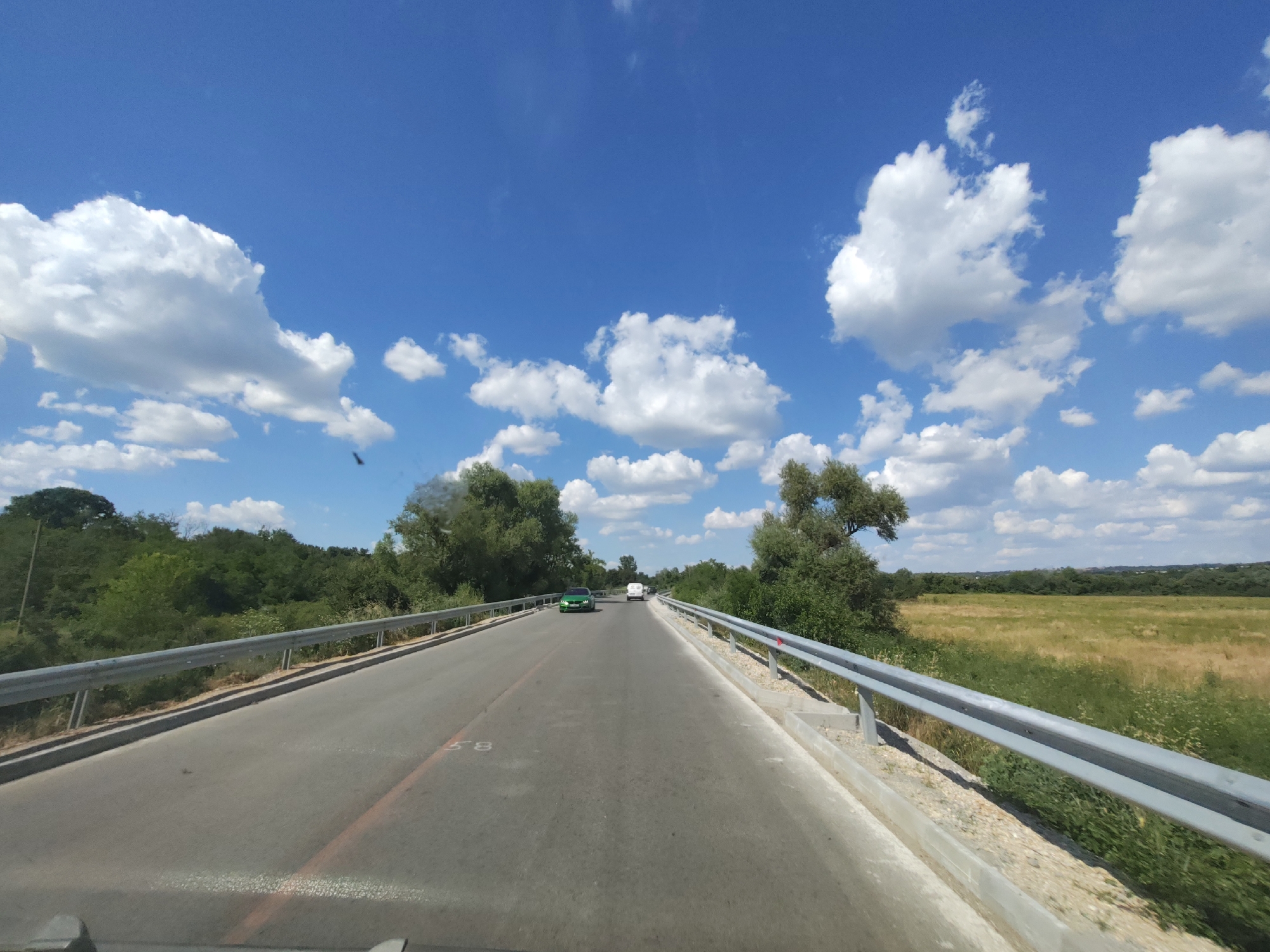 The image depicts a view from a vehicle traveling on a two-lane road that appears to be a bridge. The road has white lane markings and a red line marking on the left side, with the number "58" visible on the asphalt. Guardrails line both sides of the road.
In the distance, two vehicles are visible: a green car and a white van. The road is flanked by lush green trees and vegetation on either side, and beyond that, a wide, open field of tall grass and crops stretches towards a treeline on the horizon. The sky is a vibrant blue with numerous fluffy white cumulus clouds, suggesting a bright, sunny day. The overall scene conveys a sense of travel through a rural, possibly summery, landscape. There are no people directly visible in the image, though the presence of vehicles implies human activity. The setting appears to be Aglen, Bulgaria.