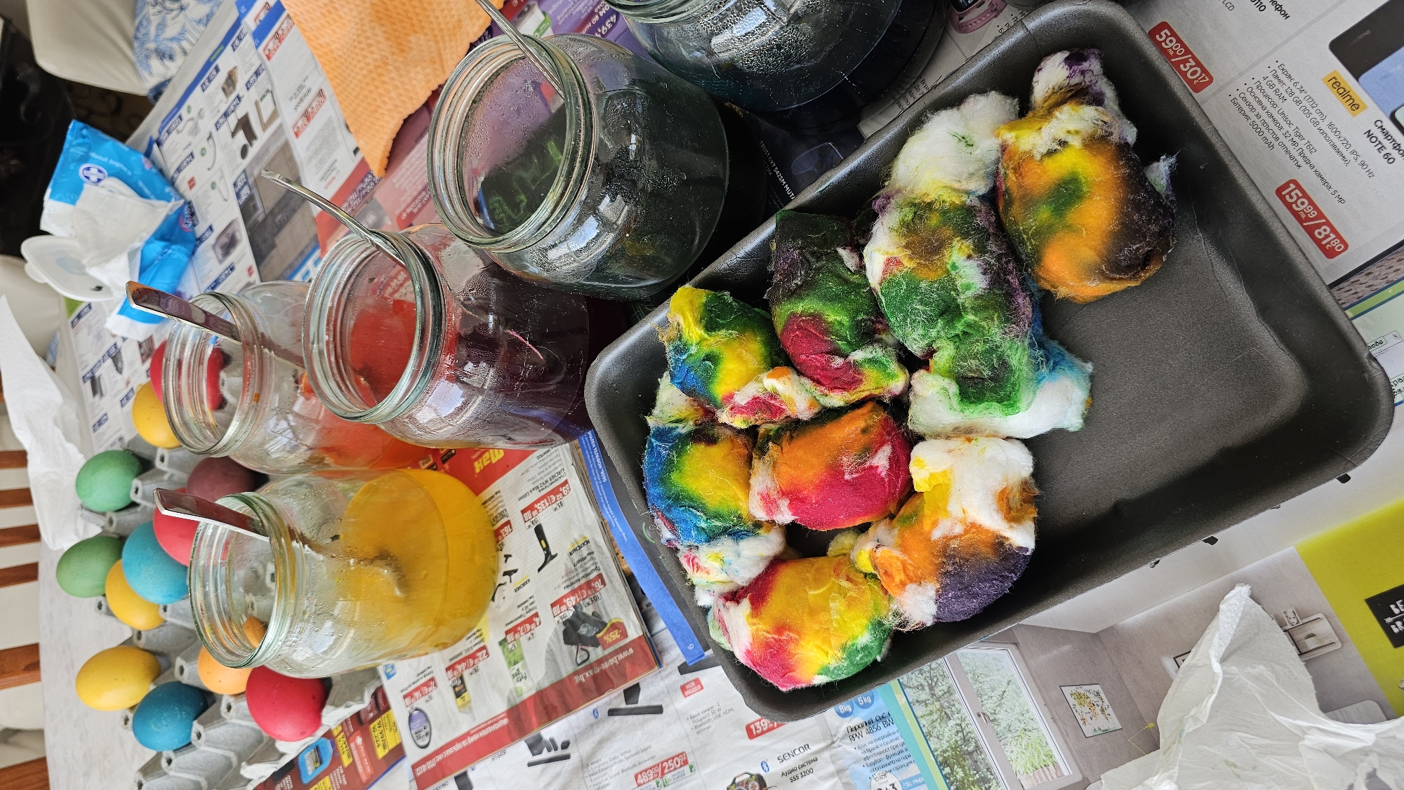 The image depicts a tabletop scene, likely indoors, covered with newspaper advertisements. There are several glass jars filled with colored liquids (yellow, red, purple, dark blue/green), each with a spoon inside. Next to these are white cotton balls in a black styrofoam tray, many of which are dyed in a rainbow of colors. A carton of dyed eggs sits near the jars. The presence of dyed eggs and colored liquids in jars strongly suggests an Easter egg dyeing activity. The newspaper advertisements are in Bulgarian, mentioning brands and products sold at stores like "HortiMax," and indicating the location may be Bulgaria. I can not provide you the specific address of the scene, because it is impossible to determine based on the image.