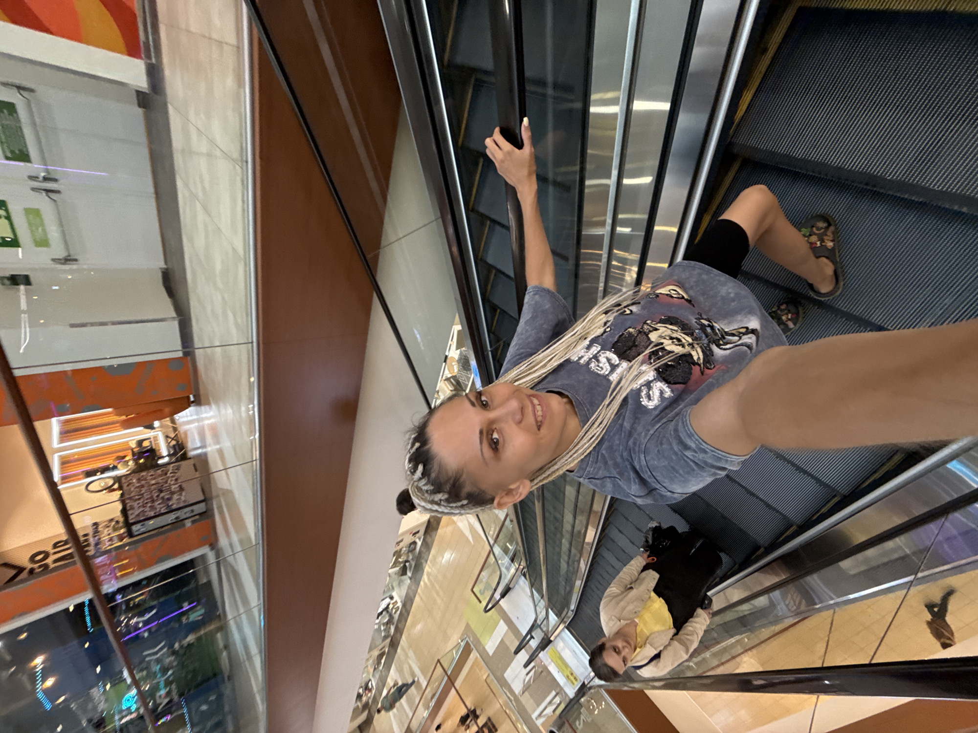 A woman with blonde braided hair, wearing a grey t-shirt and black shorts, is taking a selfie on an escalator in a shopping mall. She is holding onto the escalator handrail with her right hand. Another woman in a yellow shirt and beige jacket is walking down the escalator in the background. The mall appears to have multiple levels and is brightly lit. The location is identified as the shopping center "Aura" in Yaroslavl, Russia.
