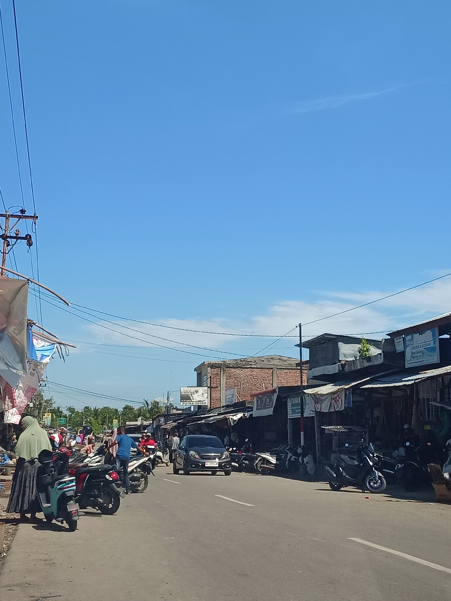 In the city of Deah Pangwa, Indonesia, a street scene unfolds under a clear blue sky with a few wispy clouds. The road is paved with asphalt and marked with white lines. On the right side of the road, a row of shops with corrugated metal roofs are visible, some displaying signs, including one that reads "PUTRA DAYAH." Several motorcycles are parked in front of these shops, and a dark grey car is driving on the road. On the left side of the road, a woman in a light green hijab stands near several parked motorcycles, one of which is a teal scooter with black trim. More motorcycles and people are present further down the street, with some individuals appearing to be engaged in activities related to the shops or the street. Power lines and poles run along the left side of the street, with wires extending across the scene. Buildings with brick facades are also visible in the background. The overall impression is of a bustling commercial area in a tropical or subtropical climate.
