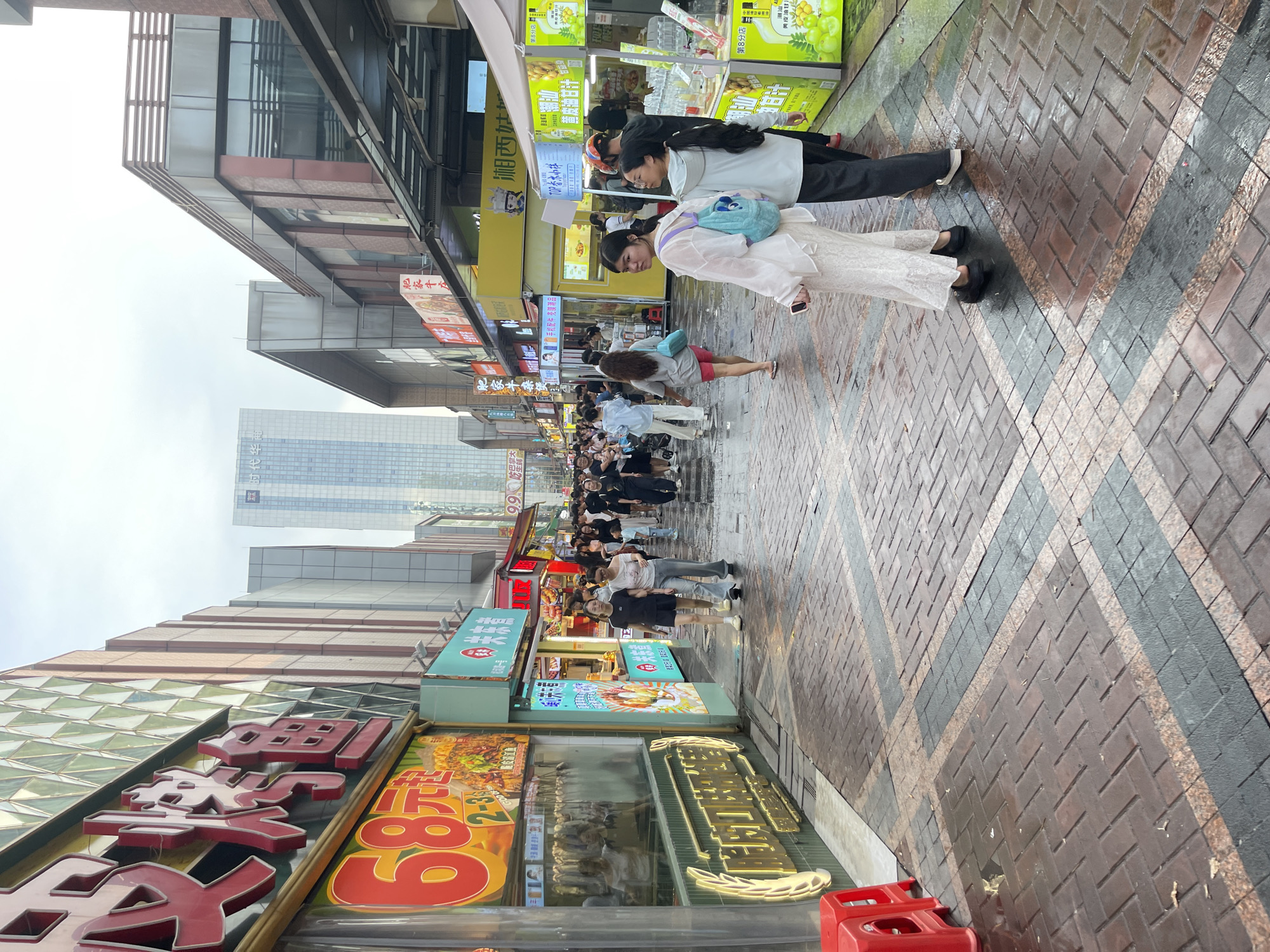 The image depicts a street scene in Panyu District, China, likely a bustling commercial area. The street is paved with dark reddish-brown and grey bricks arranged in a herringbone pattern. The pavement appears wet, suggesting recent rain. On the left side of the street, a food stall is prominently displayed with a large red sign featuring Chinese characters and a prominent "68" in a circle, indicating a price or promotion. This stall displays various food items, including what appear to be skewers of meat or seafood. Further down the street, other food stalls and shops line both sides, with awnings and brightly colored signs. A tall, modern glass skyscraper is visible in the background, towering over the surrounding buildings. Several people are walking along the street, dressed in casual clothing. Two women in the foreground are walking away from the camera, one wearing a white lace dress and the other a grey hoodie. Other pedestrians are visible in the midground and background, some walking individually, others in groups. The overall atmosphere is one of a lively urban environment, likely a popular spot for shopping and dining.