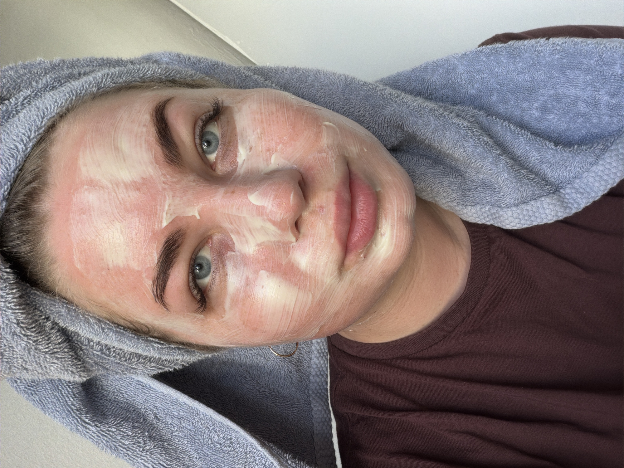 A woman with a white facial mask is seen in a close-up, portrait-style shot. She has a light blue towel wrapped around her head. The woman has fair skin, light blue eyes, and defined eyebrows. She's wearing a maroon t-shirt. There is no indication of this image being taken in Chaikovsky, Russia. The scene appears to be indoors.