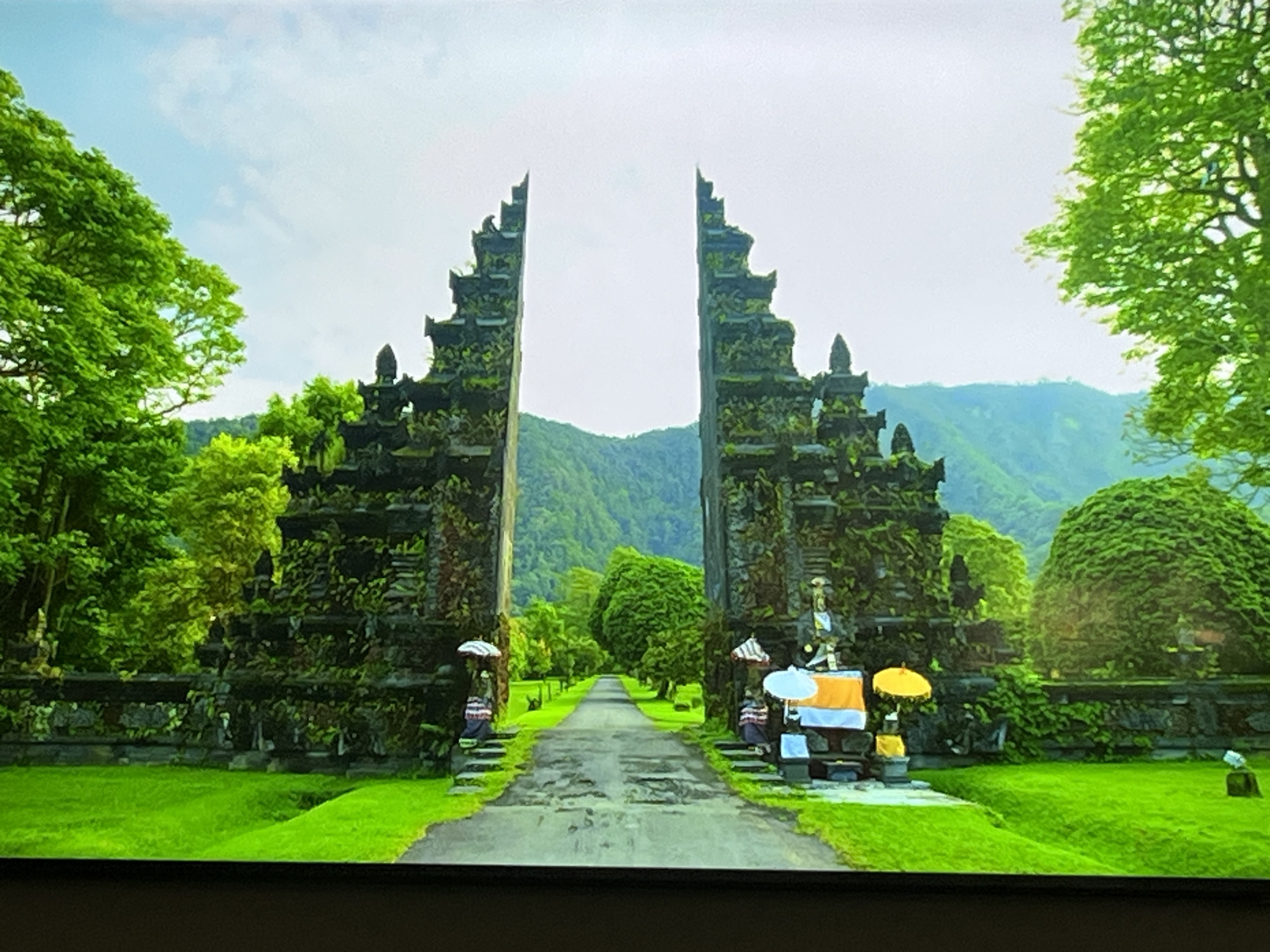 The image depicts a grand stone gateway with intricate carvings, flanked by lush green trees and a manicured lawn. The gateway is designed in a tiered architectural style, with moss and vegetation growing on its surfaces. A paved path leads through the gateway towards a backdrop of rolling green hills and a cloudy sky. Decorative umbrellas and offerings are visible near the gateway. The scene conveys a sense of tranquility and natural beauty. There is no information in the image that suggests this location is in Kharkiv, Ukraine.