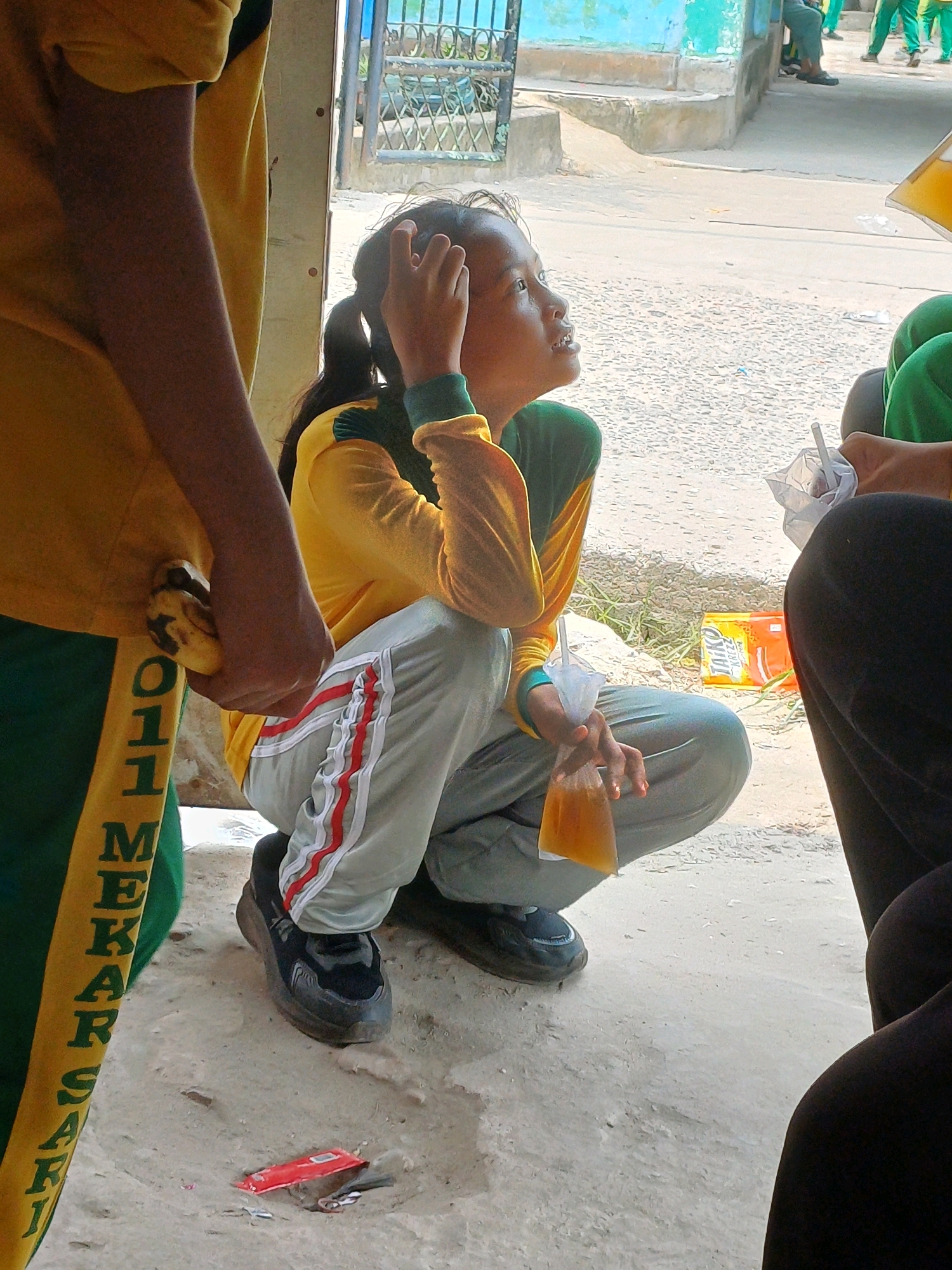In Dumai City, Indonesia, a young girl with dark hair tied in a ponytail, wearing a yellow and green long-sleeved shirt and gray pants with red stripes, sits on the ground looking upwards with her hand touching her head. She holds a plastic bag filled with a yellow liquid and a straw. Beside her, to the left, a person wearing a yellow shirt and green pants with yellow lettering on the side holds a banana in their left hand. In the background, a blue and white building with a metal fence is visible, along with other people standing. The ground appears to be sandy or dirt, with some scattered debris. An orange and yellow packaged product is on the ground near the girl.