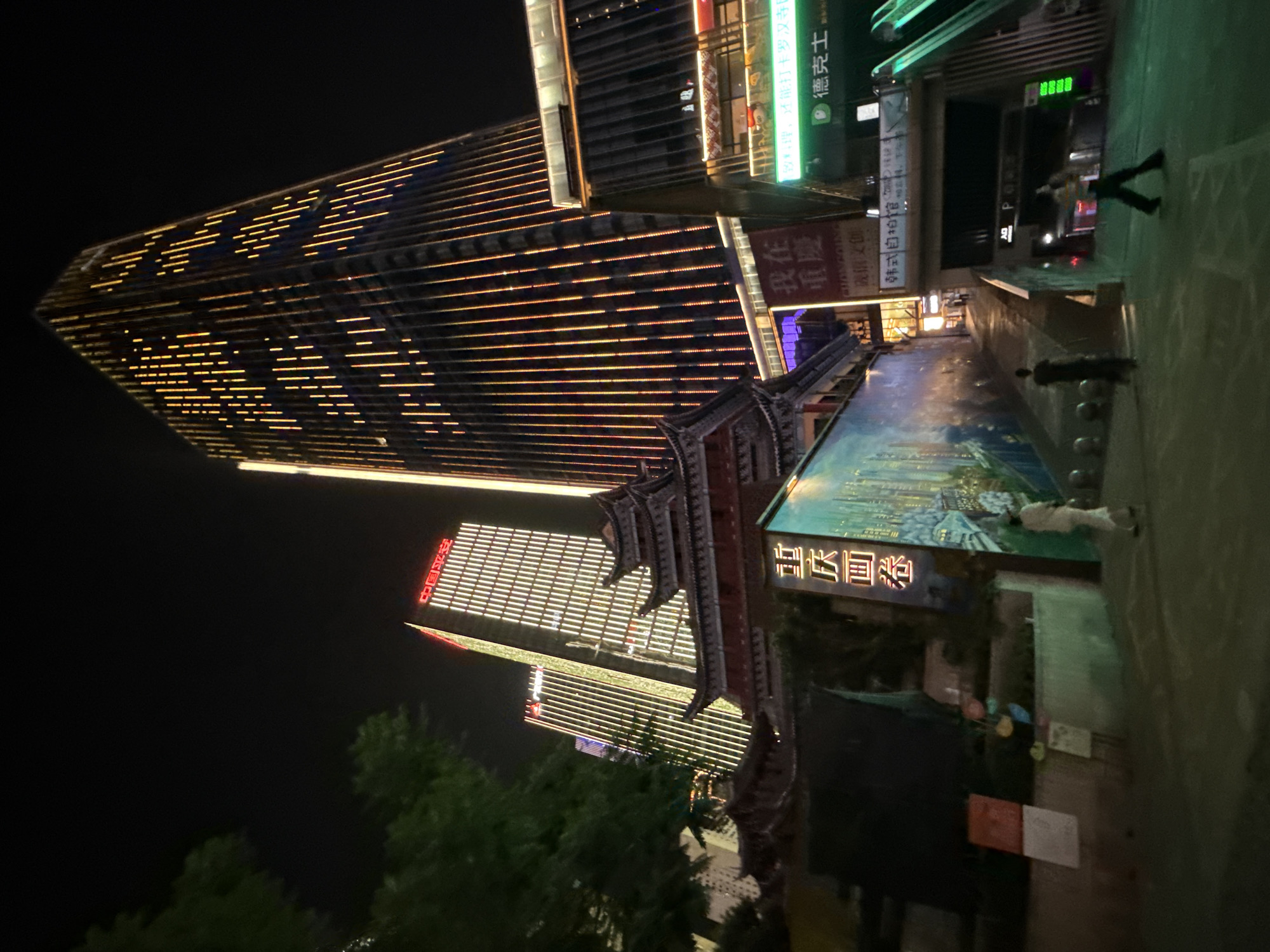 A low-angle, night-time photograph captures a busy street scene in Yuzhong District, China. Towering modern skyscrapers dominate the background, their facades illuminated by vertical streaks of orange and white lights. In the foreground, a traditional Chinese-style building with layered, dark tiled roofs stands in contrast to the contemporary architecture. A vibrant mural depicting a cityscape adorns the wall of a structure to the left, its colors glowing under the artificial light. Several people are visible in the scene. Two individuals are walking away from the camera on the right side, and another figure in a light-colored outfit is walking towards the left. The street is paved, and bollards line the walkway. An escalator leading underground is visible on the far right, with green illuminated signage. The overall impression is one of a dynamic urban environment where modern and traditional elements coexist.