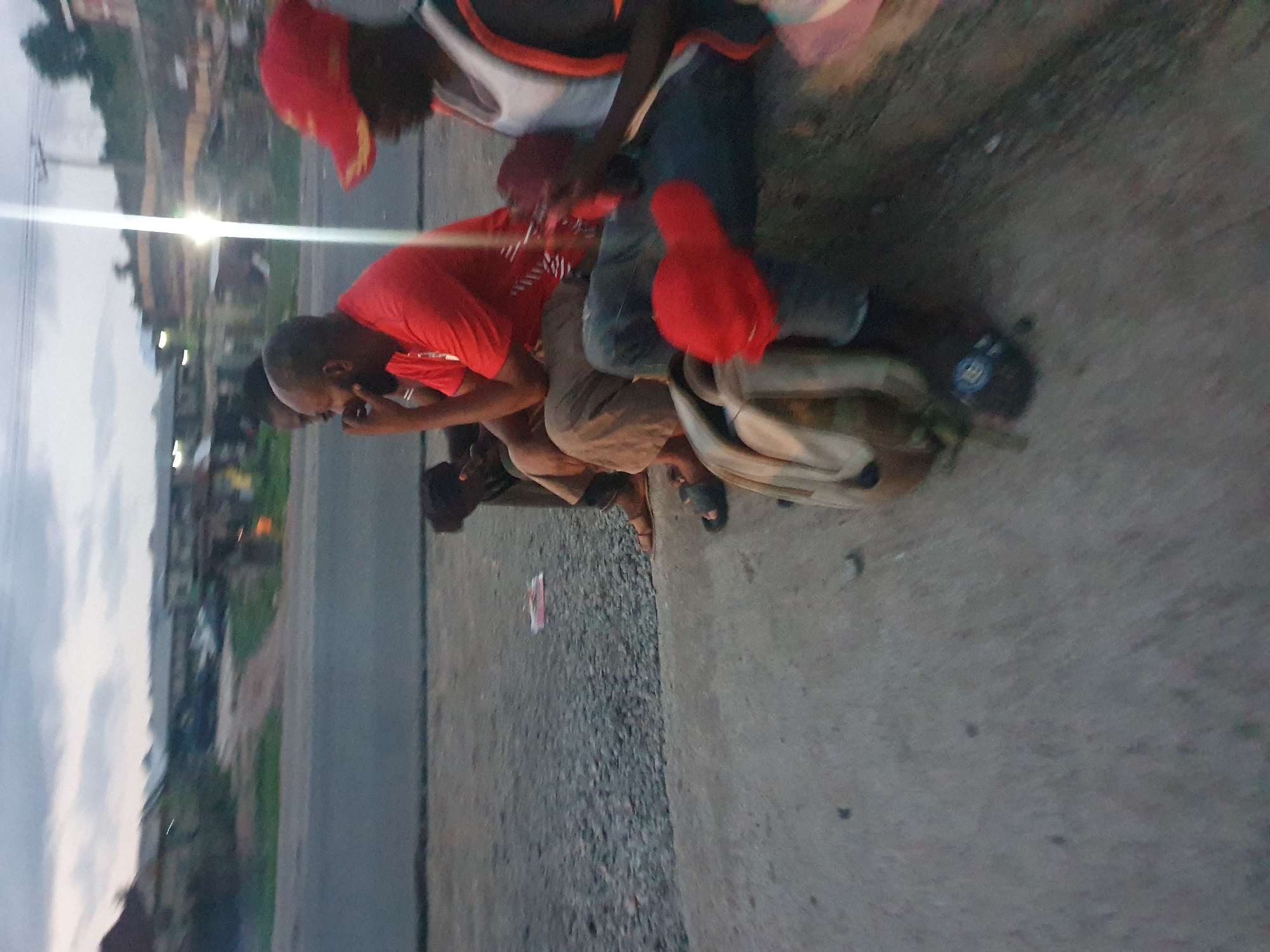The image shows three or four dark-skinned males seated on a paved surface in Lokoja, Nigeria. The individuals are wearing red T-shirts and at least two are wearing red caps. One male, closest to the camera, is dark-skinned, bald, and appears to be using a cell phone. A fourth individual can be seen to his left in the background. There's a tan backpack on the ground next to them. The background shows a street and buildings in the distance. It appears to be dusk or evening.