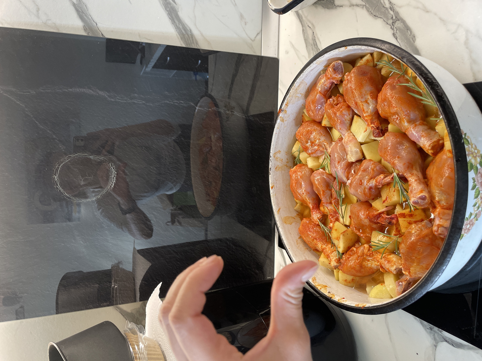 The image shows a cooking scene on a kitchen countertop. A black induction cooktop sits beside a white countertop with marble-like patterns. A large, white enamel cooking pot is placed on the cooktop, filled with seasoned chicken drumsticks and diced potatoes, garnished with rosemary sprigs. A hand is partially visible in the foreground, holding a container with white paper towels inside. The reflection in the cooktop shows a person holding a phone, capturing the image. The scene appears to be indoors, likely in a domestic kitchen. The exact location in Sofia, Bulgaria, cannot be determined from the image alone.