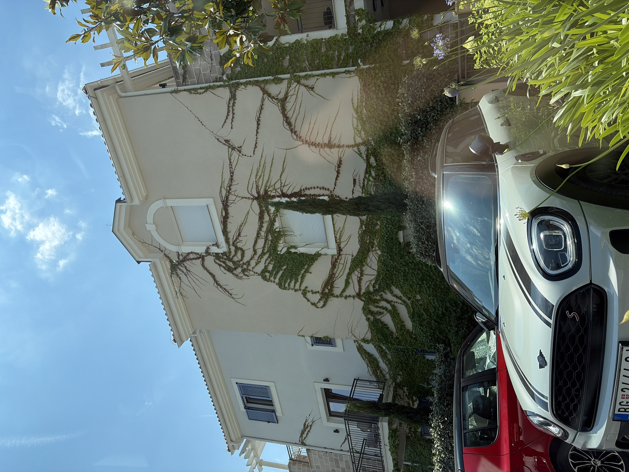 The image depicts a sunny outdoor scene in Radovići, Montenegro. In the foreground, a light-colored Mini Cooper is parked, with its hood and front bumper prominently visible, featuring black racing stripes. To the left of the Mini Cooper, a red hatchback is partially visible. The background showcases a light-colored, multi-story building with climbing ivy covering a significant portion of its facade. Windows with dark shutters and a balcony are also visible on the building. Lush green foliage and some flowering plants are present in the lower right portion of the frame. The sky is clear blue with scattered white clouds. The scene appears to be a residential area or a property associated with a hotel or resort.