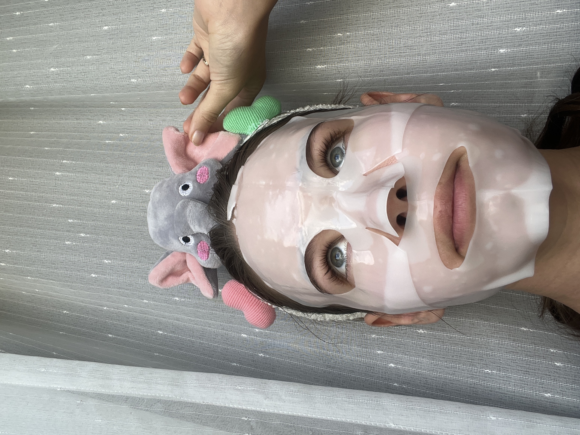 A person wearing a white facial sheet mask and an elephant-shaped headband is lying on a striped gray surface. A hand is partially visible above the person's head. The location is not discernible from the image.