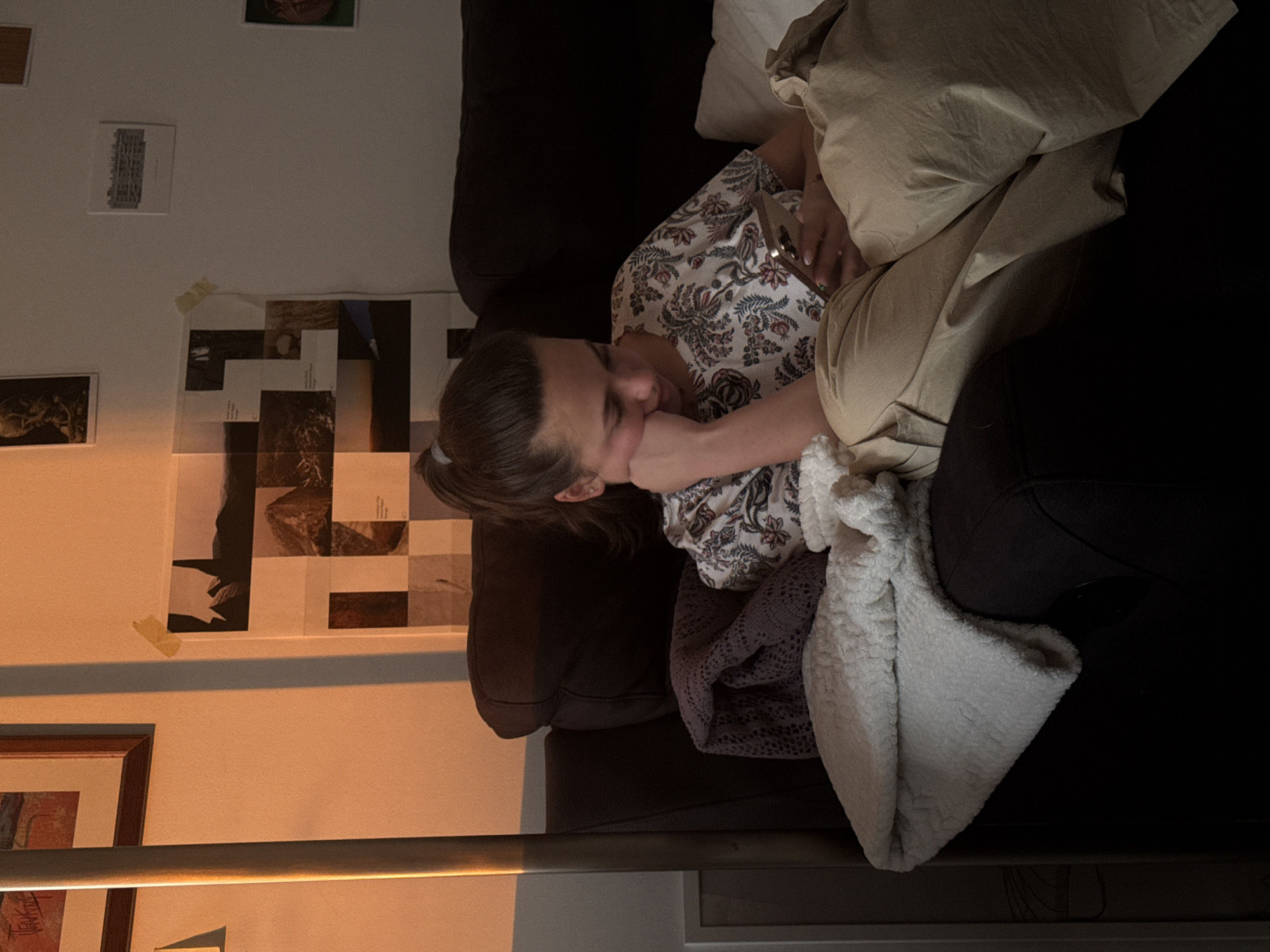 A young person with brown hair pulled back in a ponytail is lying on a dark brown sofa. They are wearing a floral print shirt and tan pants. They are resting their face in their hand and holding a cell phone. A tan pillow and a white blanket are beside them. The wall behind the sofa is decorated with several framed pictures and a grid of taped images. The lighting is warm, suggesting it might be evening. There is no information available to determine if the photo was taken in Milan, Italy.
