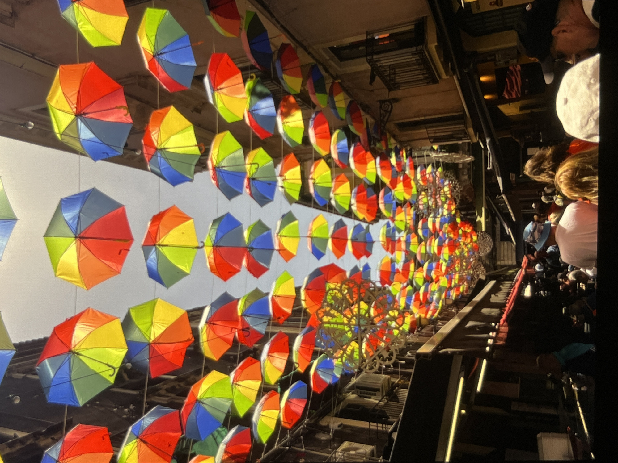 An overhead shot captures a vibrant street scene where numerous colorful umbrellas are suspended above, creating a canopy of red, yellow, green, blue, and orange. The umbrellas are arranged in a dense, staggered pattern, casting a kaleidoscope of colors onto the street below. Buildings with balconies line both sides of the narrow street, some with lights on, suggesting shops or residences. The sky above is overcast. In the lower portion of the image, people are visible walking along the street, their faces turned away from the camera or obscured. They are dressed in casual clothing, and some are wearing hats. The overall impression is of a lively, festive urban environment decorated for an event or as a permanent artistic installation.