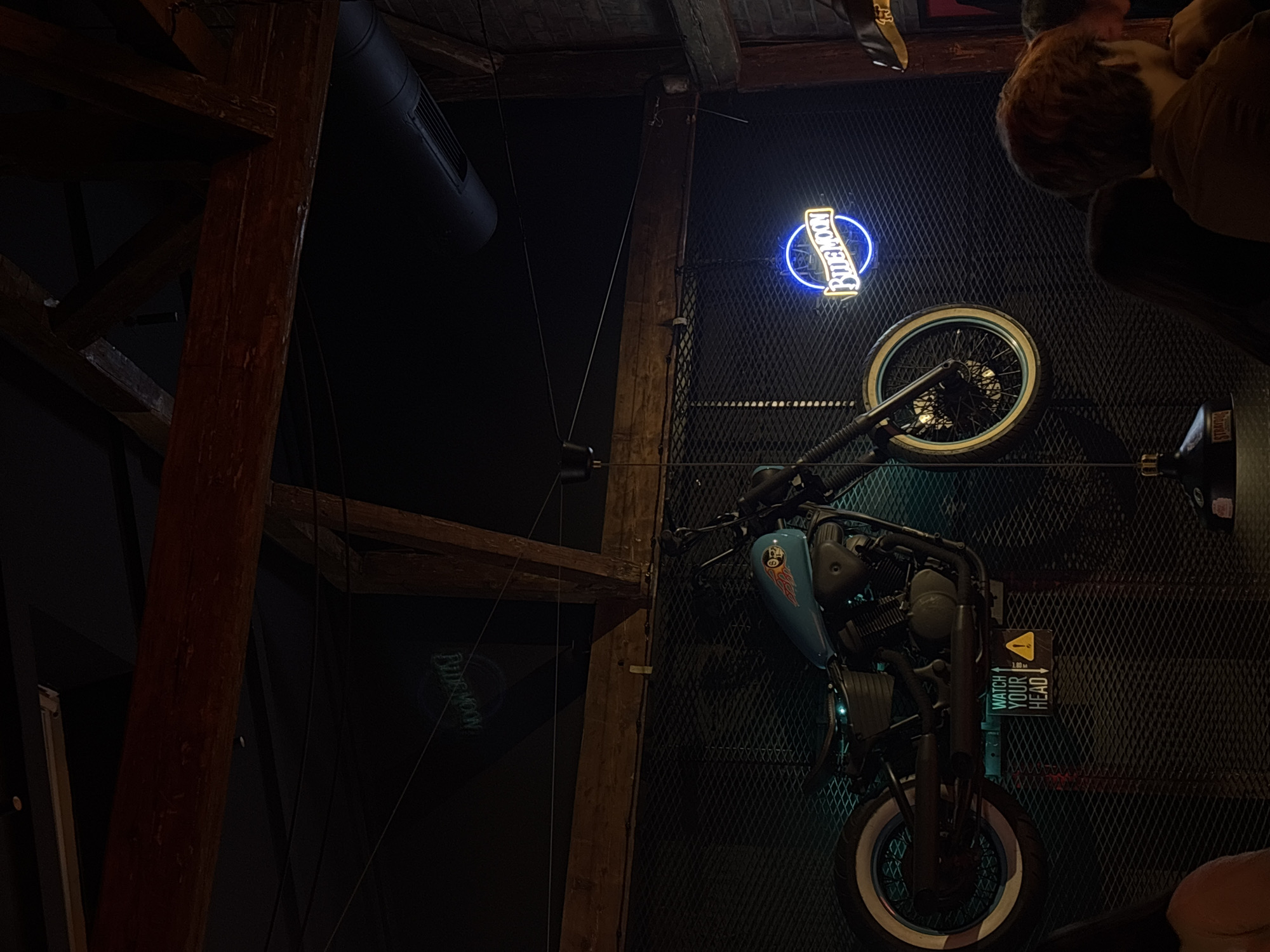The image shows a bar or restaurant interior with a decorative motorcycle mounted on a wire mesh wall. The motorcycle is light blue and black with white-walled tires. A "Blue Moon" neon sign is mounted above the motorcycle's rear tire. A "Watch Your Head" sign is mounted near the front of the motorcycle. The interior design features dark wood beams and a black ceiling with a ventilation pipe. A person is partially visible in the upper right corner. The location is a bar or restaurant, potentially in Belgrade, Serbia based on the "Blue Moon" sign.