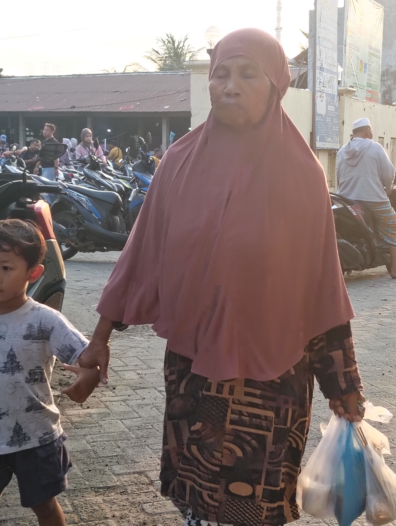 In Banjarwati, Indonesia, an elderly woman wearing a maroon hijab and a patterned dress walks with a young boy. The woman holds two plastic bags filled with groceries in her right hand. The boy, on the left, wears a t-shirt and shorts and holds the woman's left hand. They are on a paved area, possibly a market or roadside, with numerous motorcycles parked in the background. A building with a corrugated metal roof is visible behind the motorcycles. The lighting suggests it is either early morning or late afternoon.