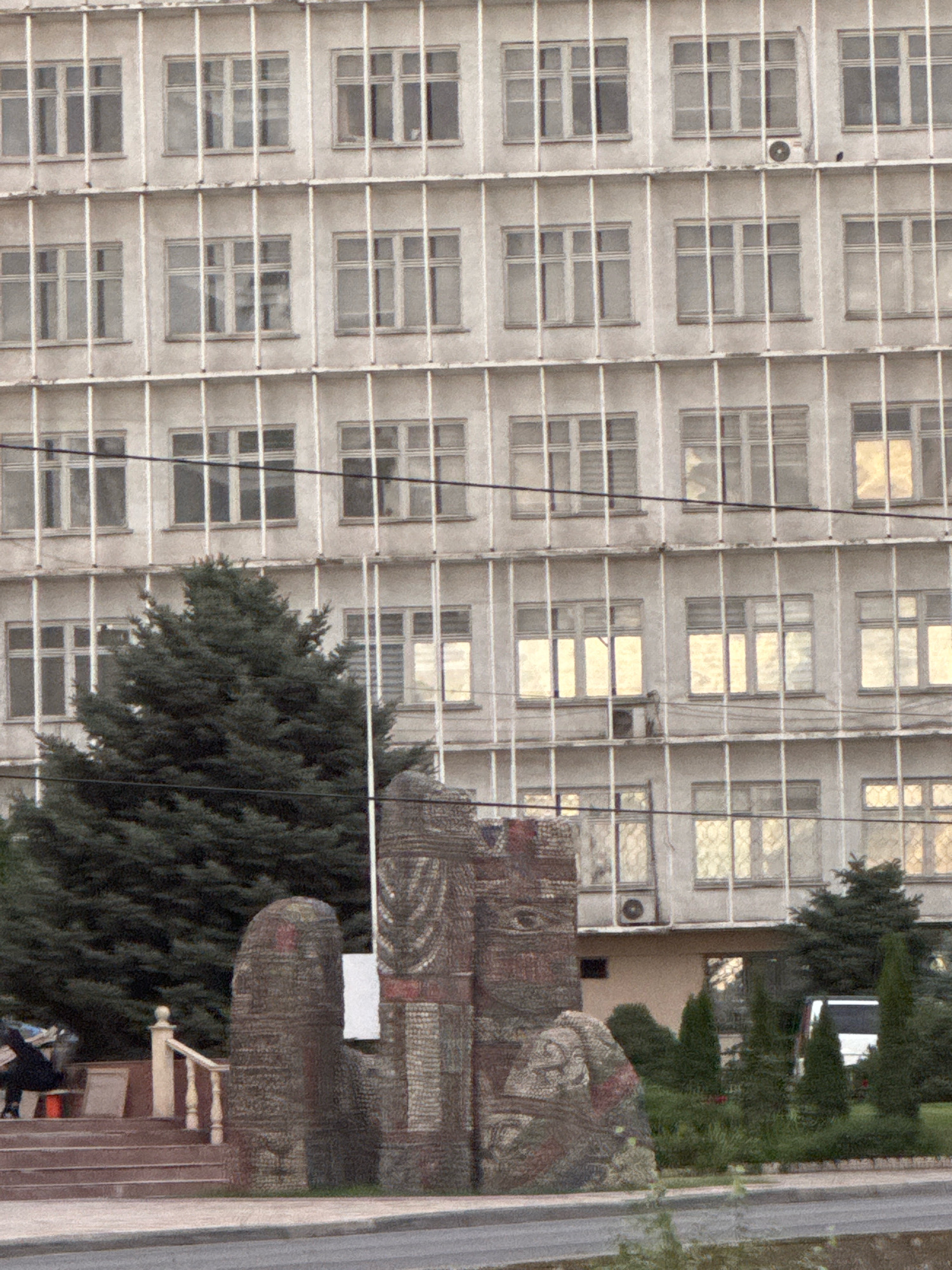 The image shows the facade of a Brutalist-style building with a grid of windows. In the foreground, there are several abstract stone sculptures and a large evergreen tree. To the right, a white minivan is parked near a row of small trees. The building appears to be old, with some visible signs of wear and tear. The overall scene suggests an outdoor urban environment in Bishkek City, Kyrgyzstan. There are no visible people in the image.