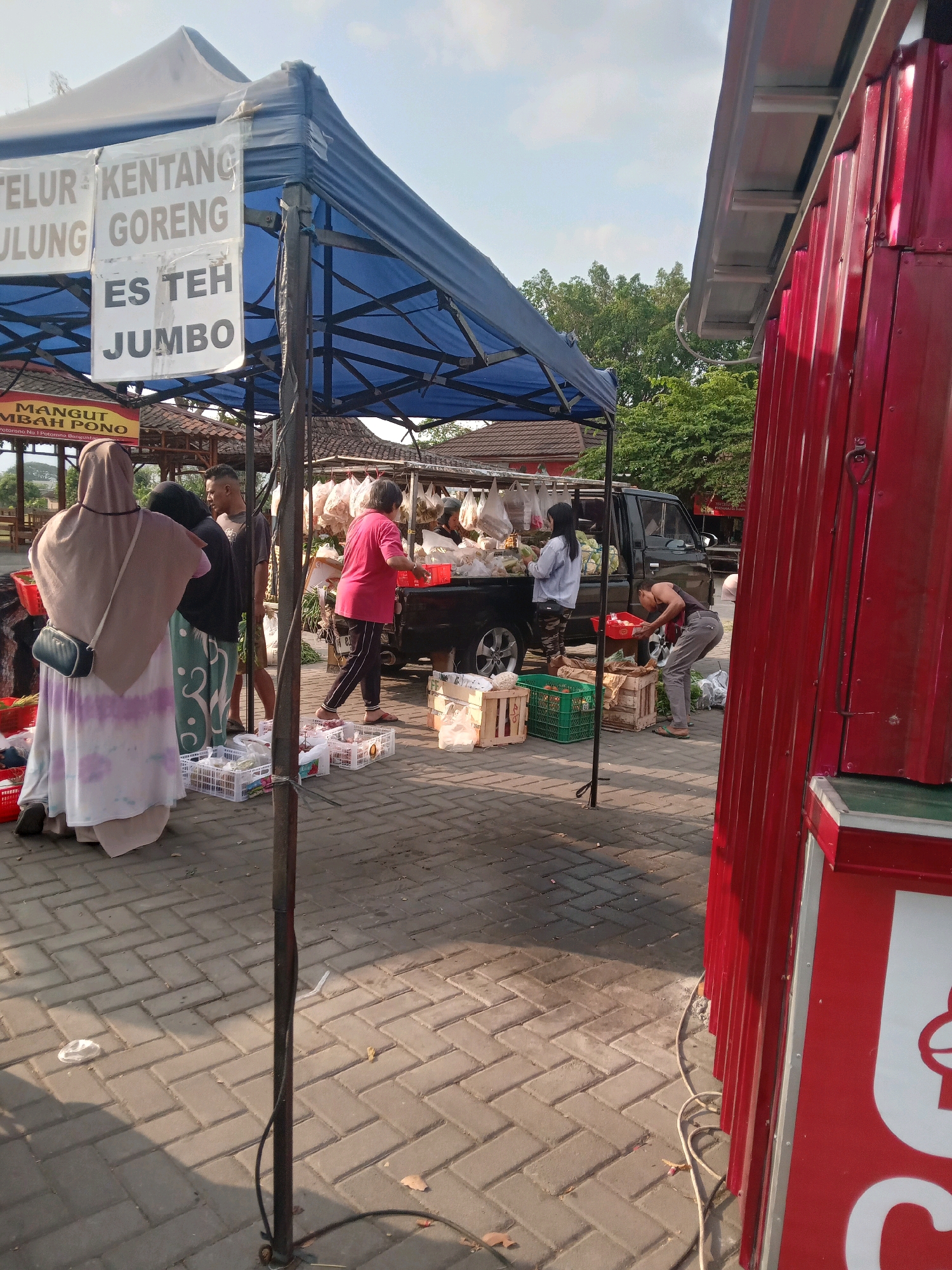 The image depicts an outdoor market scene in Banguntapan, Indonesia, with a focus on food stalls. A blue tarp-covered stall displays signs in Indonesian, including "TELUR KENTANG GORENG" (Fried Potato Eggs) and "ES TEH JUMBO" (Jumbo Iced Tea), indicating food and beverage offerings. Several people are present, including a woman in a headscarf and a patterned skirt standing near the stall, and others interacting with goods at various stands. In the background, a black pickup truck is parked, and more stalls with hanging produce are visible. To the right, a bright red corrugated metal structure, likely another stall or shop, dominates the foreground. The ground is paved with interlocking brick pavers. The lighting suggests it is daytime, with shadows cast across the scene.