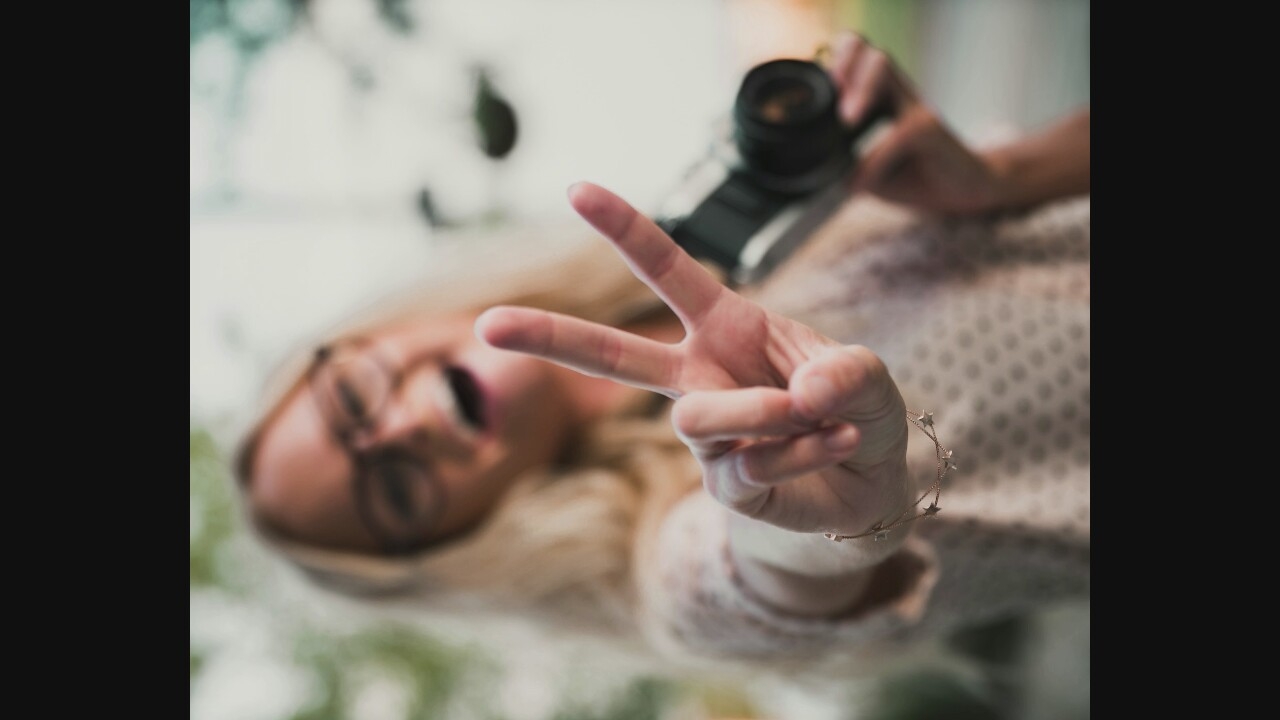 In an outdoor, blurred setting, a woman with blonde hair and glasses is captured from the chest up, holding a vintage video camera in her right hand and making a peace sign with her left hand. She is wearing a light-colored top with a subtle pattern. The background is out of focus, with hints of greenery suggesting an outdoor environment.