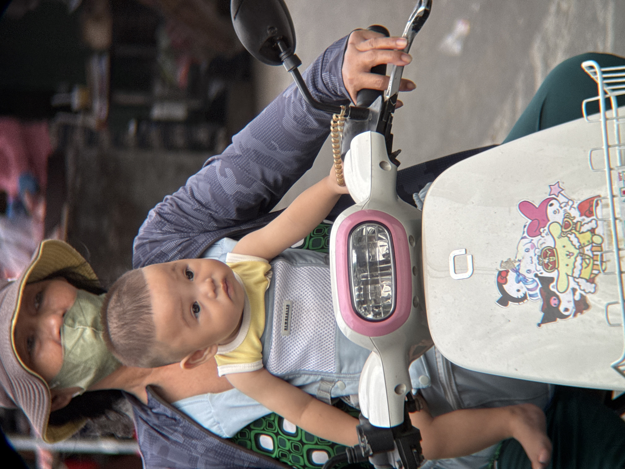 The image shows a person riding a scooter in what appears to be a city environment, likely Nha Trang, Vietnam, based on visual cues. The rider, a woman wearing a wide-brimmed hat and a light green face mask, is seen from the chest up, with her face partially obscured by the mask and hat. She is wearing a dark, patterned jacket over a light blue garment and a green top with a white and green geometric pattern. A young child is secured in a baby carrier on the rider's chest. The child is wearing a yellow and white tank top. The scooter's handlebar area is visible, including a pink-trimmed headlight and decorative stickers featuring cartoon characters on the front panel. The background is blurred but suggests a street scene with buildings and possibly other vehicles. The rider's hand is on the scooter's handlebar. The overall lighting suggests an outdoor daytime setting.