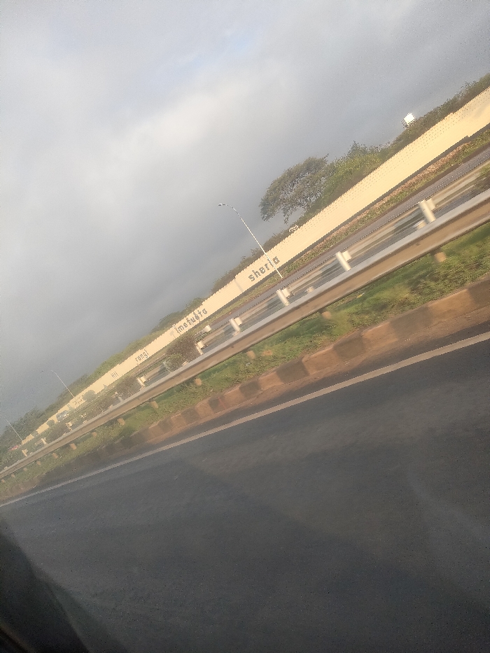 This image presents a diagonally oriented, matter-of-fact view of a multi-lane highway in Theta ward, Kenya, under an overcast sky. The foreground consists of a dark asphalt road surface, appearing slightly reflective. To the right, a brown concrete curb separates the asphalt from a strip of green grass. Beyond this, a silver-colored metal guardrail system runs parallel to the road. Behind the guardrail, another band of green vegetation precedes a tall, light-colored concrete barrier wall.
Prominently displayed on the concrete barrier, painted in black capital letters, are the Swahili words: "sheria," "Imefuta," "Rangi," and "Hii." Spaced along the wall are multiple streetlights with curved arms extending over the carriageway. In the background, indistinct vehicles are visible on an opposing carriageway behind the guardrail and wall. Sparse green trees and vegetation are visible above the concrete barrier, silhouetted against the grey sky. No individuals are discernible within the scene. The overall perspective, including reflections in the lower left, indicates the photograph was taken from inside a moving vehicle.