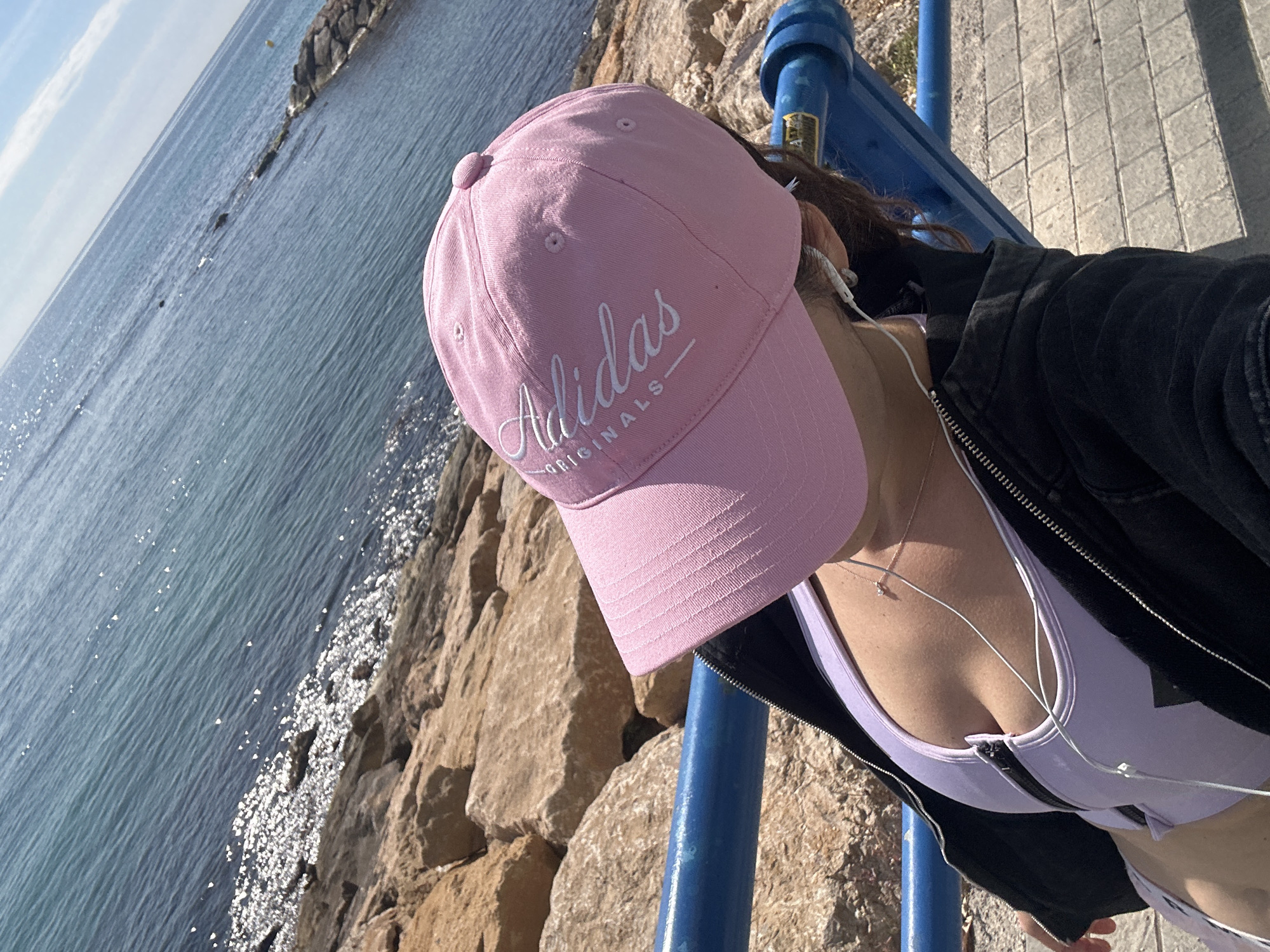 A selfie shows a person in a pink Adidas baseball cap, wearing a light purple top and a black jacket, standing near a rocky shoreline and a blue metal railing. The person is also wearing white wired headphones and a necklace with a pendant. The background features the sea and a rocky breakwater. The exact location and city are unknown.