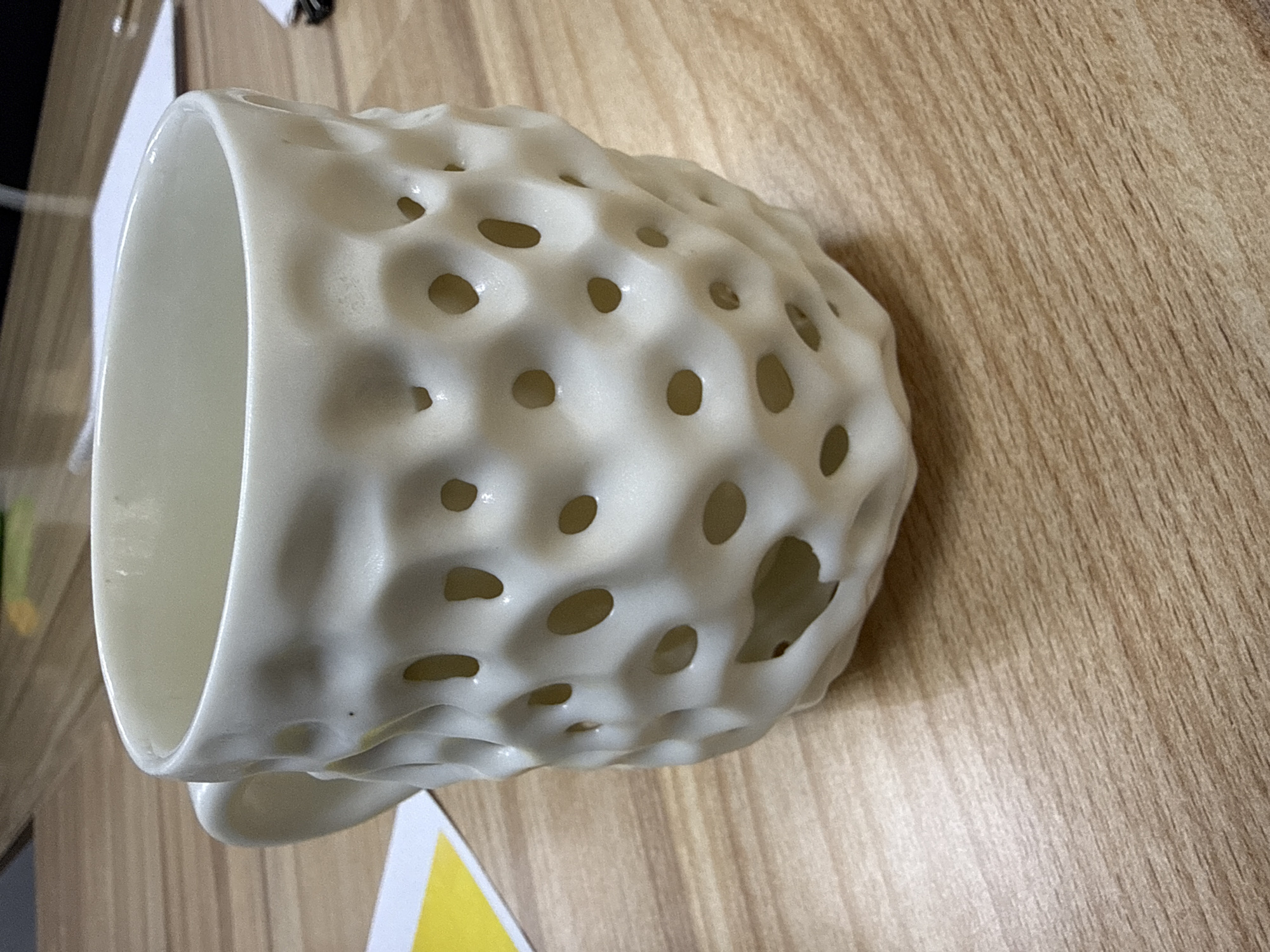 A light-colored, porous ceramic mug with rounded holes is positioned on a light brown wooden surface with a linear grain pattern. The mug is oriented at an angle, showing its interior and a small handle. A yellow and white paper object is partially visible at the bottom left of the frame. The scene is brightly lit, showing details in the mug's texture. There is no evidence of a location in Krasnoyarsk, Russia, or any people.