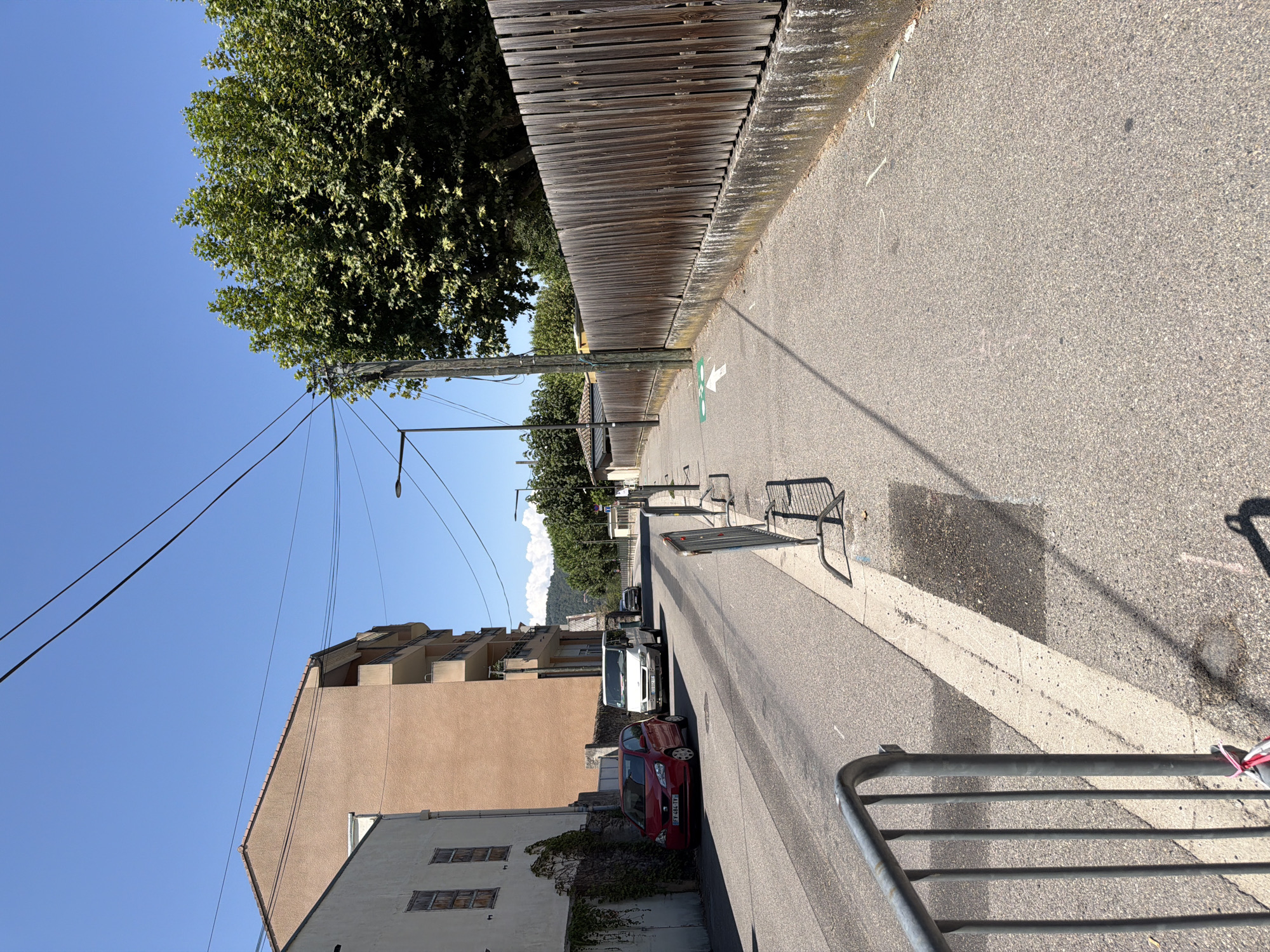 This image depicts a street scene in Tournon-sur-Rhône, France. A red car and a white van are parked on the left side of the street. Further down the street, another vehicle is visible. On the right side of the street, a wooden fence runs along a sidewalk. A green arrow painted on the sidewalk indicates a bicycle path. Overhead, power lines crisscross the blue sky. The environment appears to be an urban or suburban setting with buildings and trees present. The lighting suggests it is a sunny day.