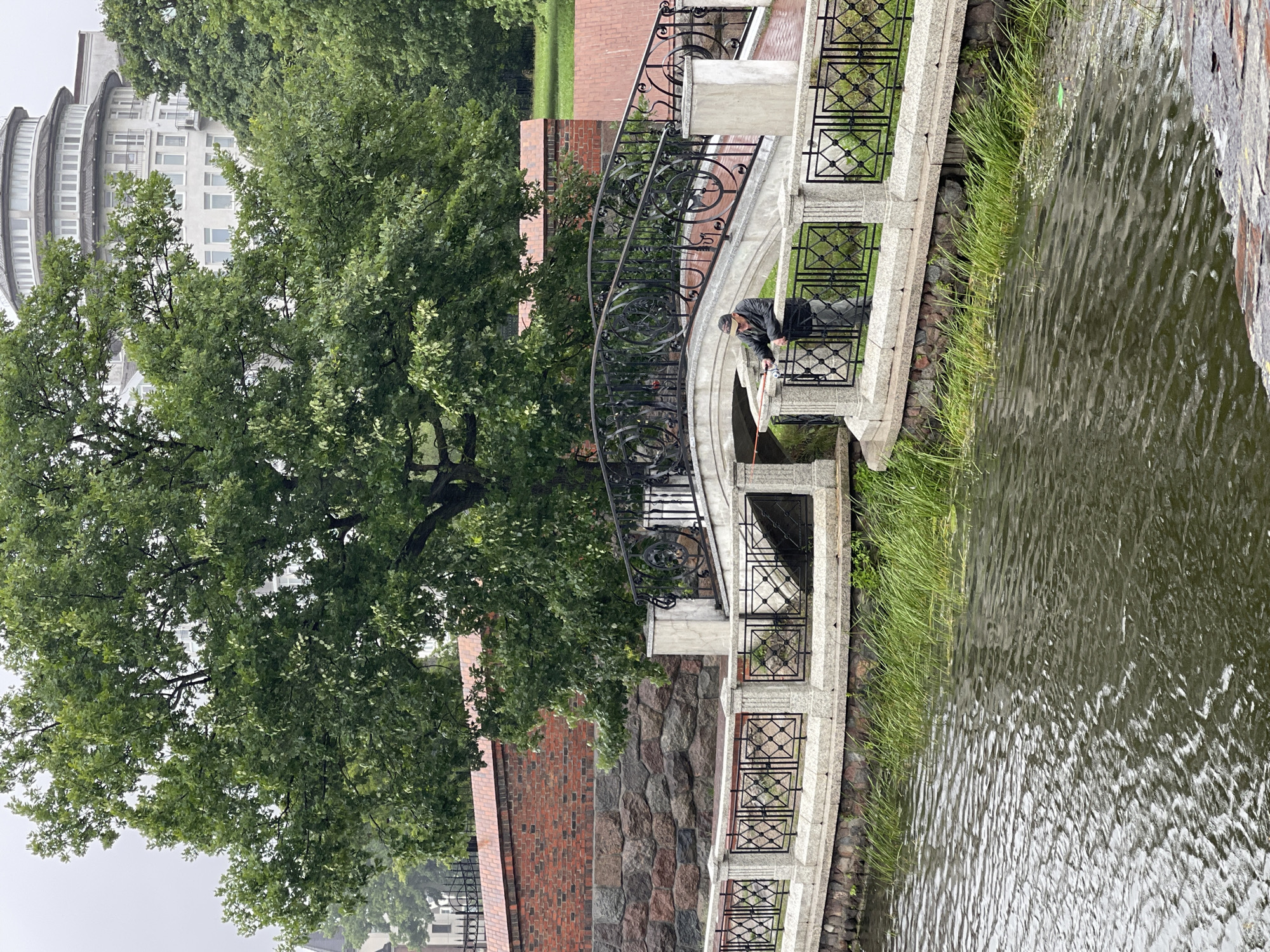 A man is fishing from a bridge in Kaliningrad, Russia. The man is wearing a dark jacket and cap, and he is holding a fishing rod. The bridge is made of stone and has a black metal railing. The bridge spans a canal, and the water is dark green. There are trees and buildings in the background. The sky is overcast. The man is standing on the right side of the bridge, and the fishing rod is in the water on the left side. The man is looking at the fishing rod. The image is taken from a low angle, and the bridge is in the foreground. The overall scene is peaceful and serene.